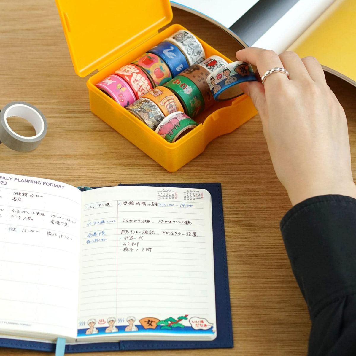 Hand lifting an illustrated washi tape roll from a yellow case, beside an open planner on a wooden desk.