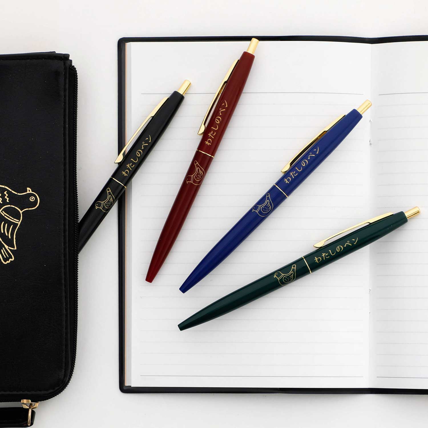 Four colored pens scattered on an open notebook beside a black zippered pen pouch.