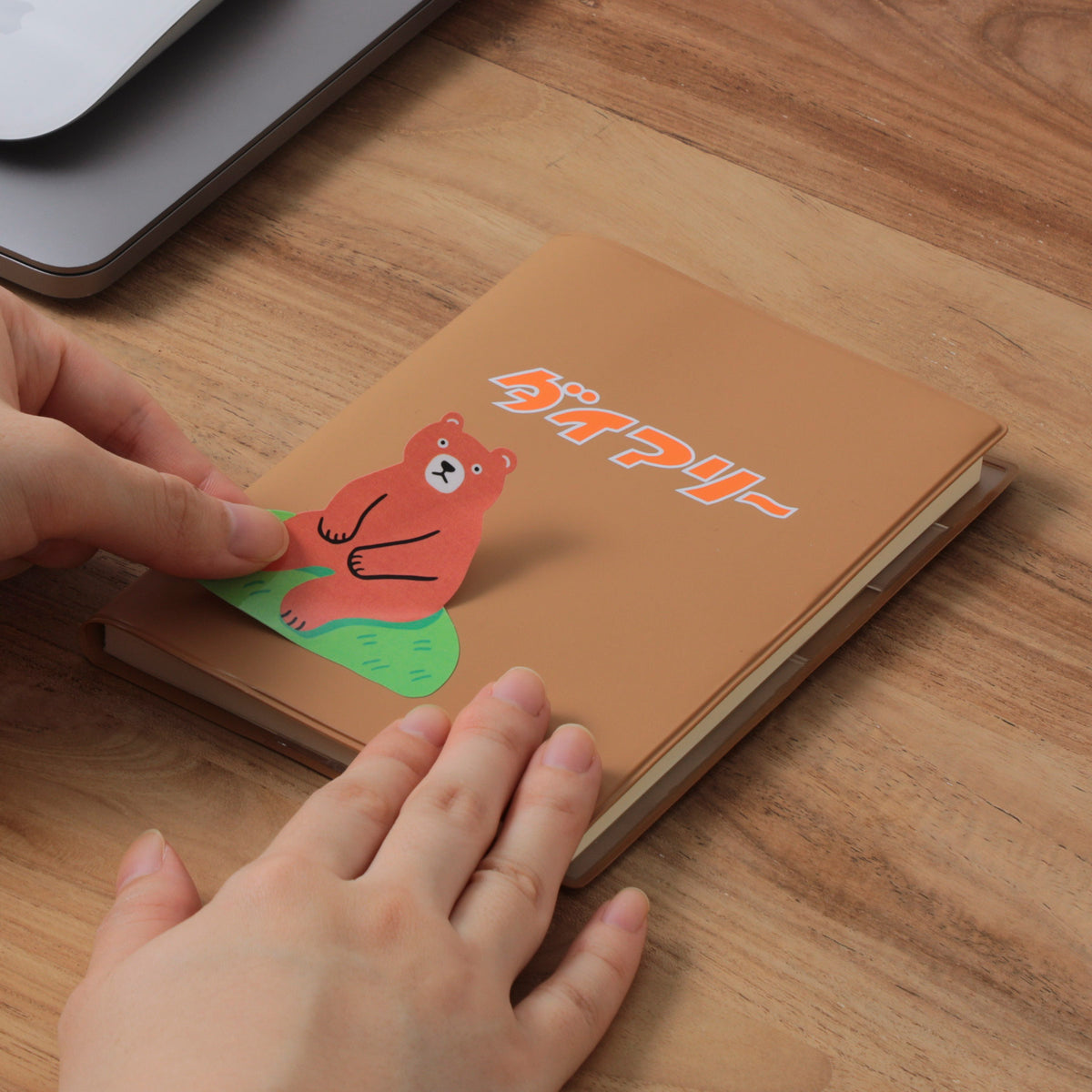 Hands applying a bear die-cut sticker to a Brown A6 planner cover on a wooden desk.