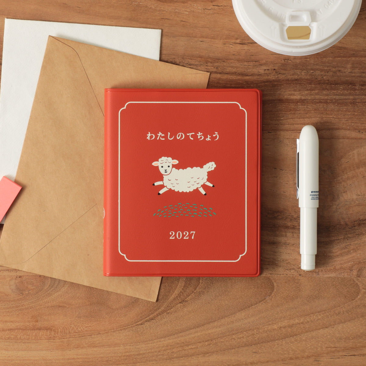 Red 2027 planner with a white sheep illustration on the cover, lying on a wooden desk beside a kraft envelope and white pen.