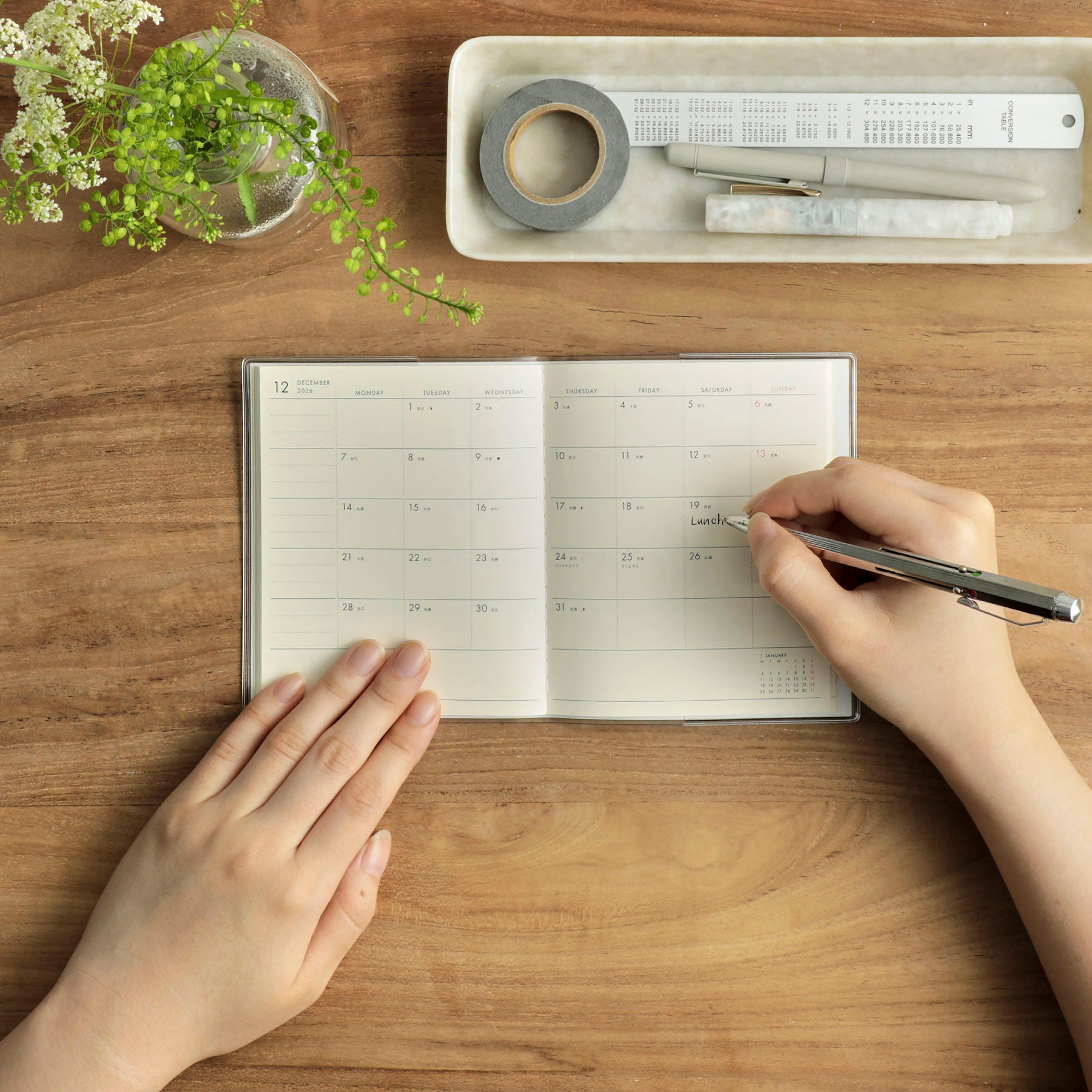 Hands writing in an open monthly planner on a wooden desk, beside a tray with pens, tape and a ruler.