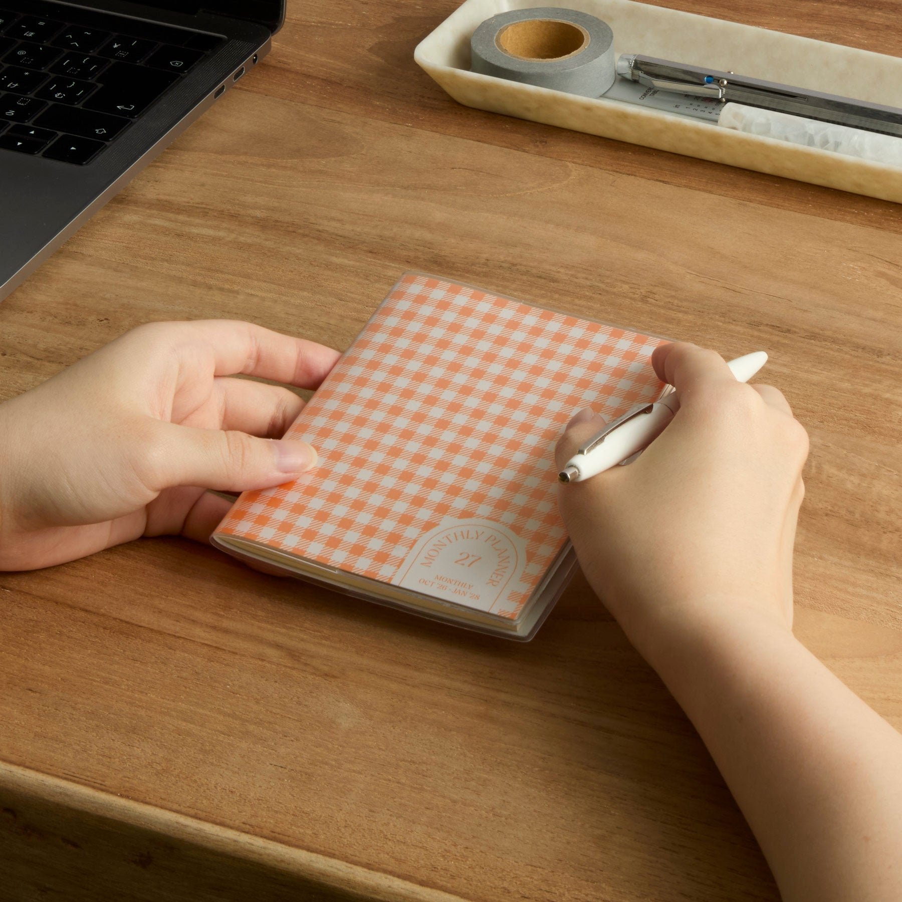 Hands holding an Orange gingham check 2027 monthly planner on a wooden desk, pen ready to write.