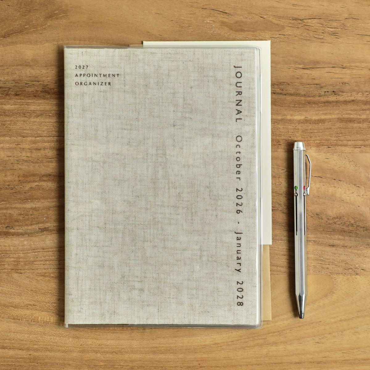 Hightide 2027 Coton A5 monthly planner in Linen with clear cover, beside a silver pen on wood.