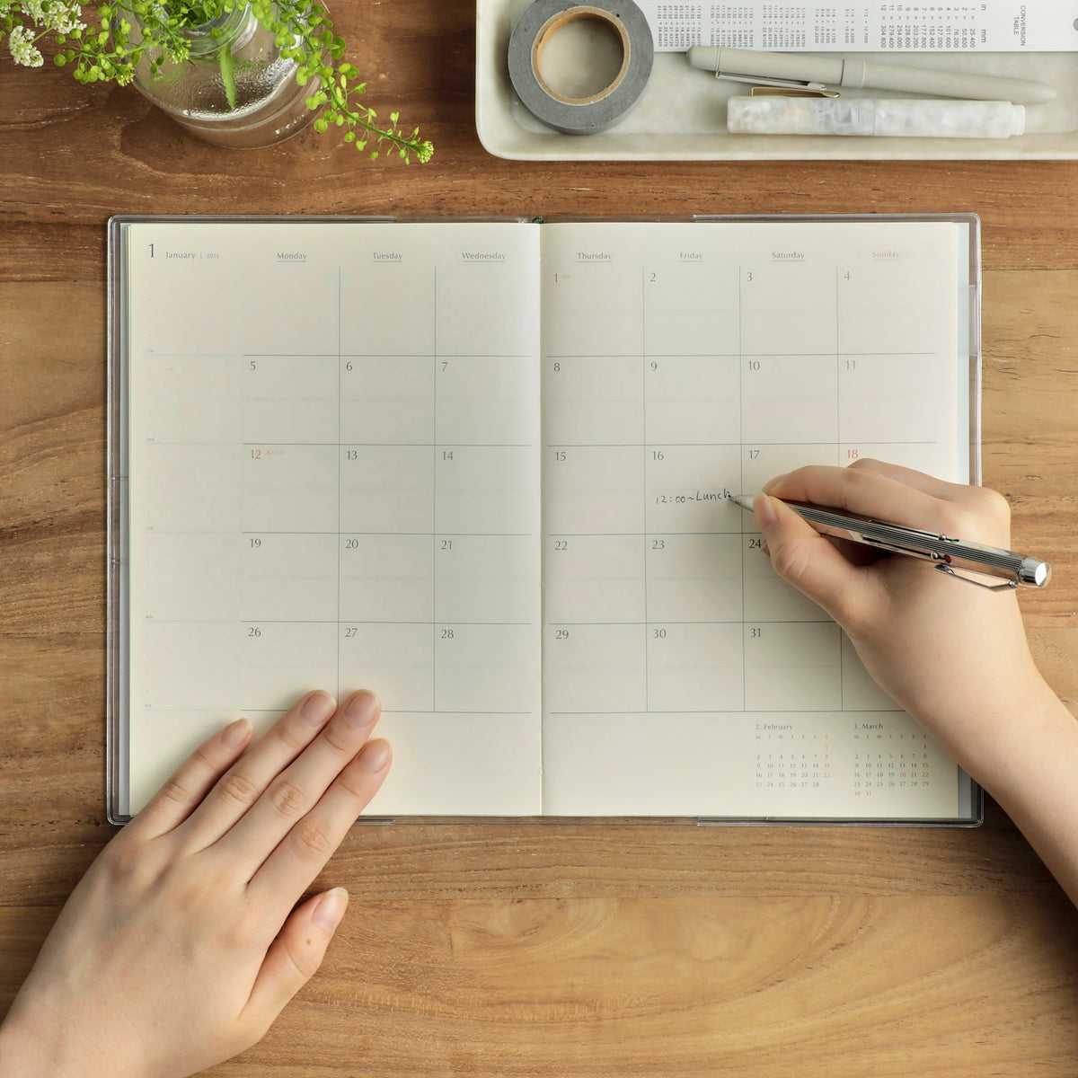 Hands writing a lunch note on the open January monthly grid of a Hightide Coton A5 planner on wood.