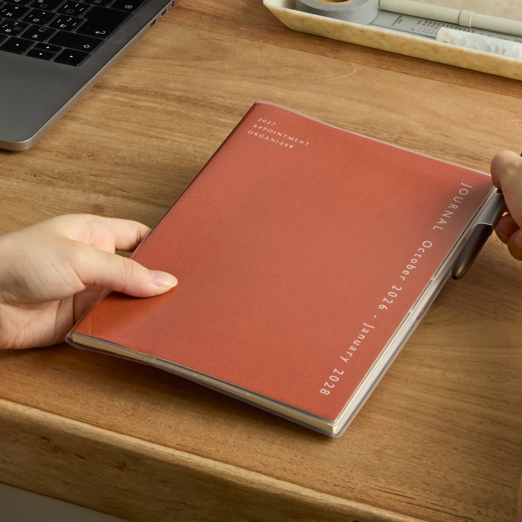 Hands holding an Orange fabric-covered Hightide planner with clear cover on a wooden desk beside a laptop.