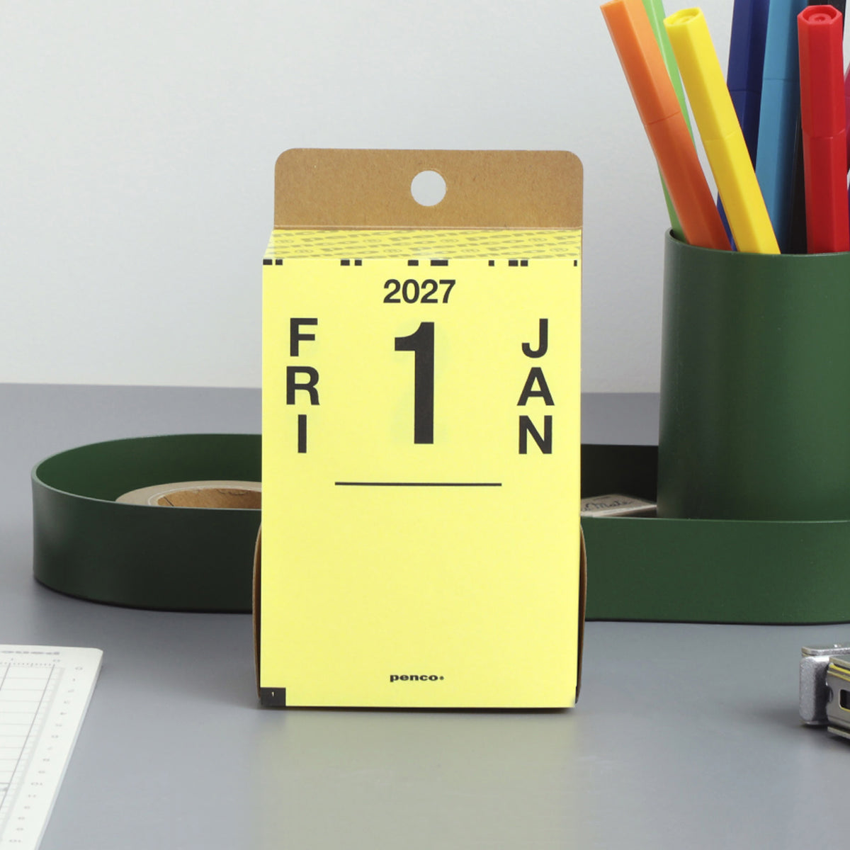 Penco 2027 daily calendar on a desk, yellow page reading FRI 1 JAN, beside a green pen cup and tray.