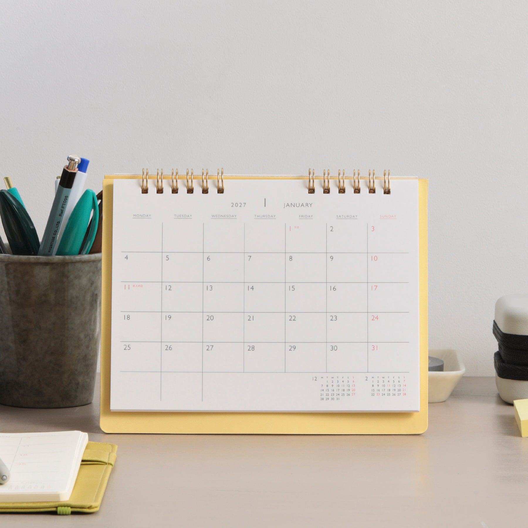 Light Yellow twin-ring desk calendar standing on a desk, open to January 2027, beside a pen cup.
