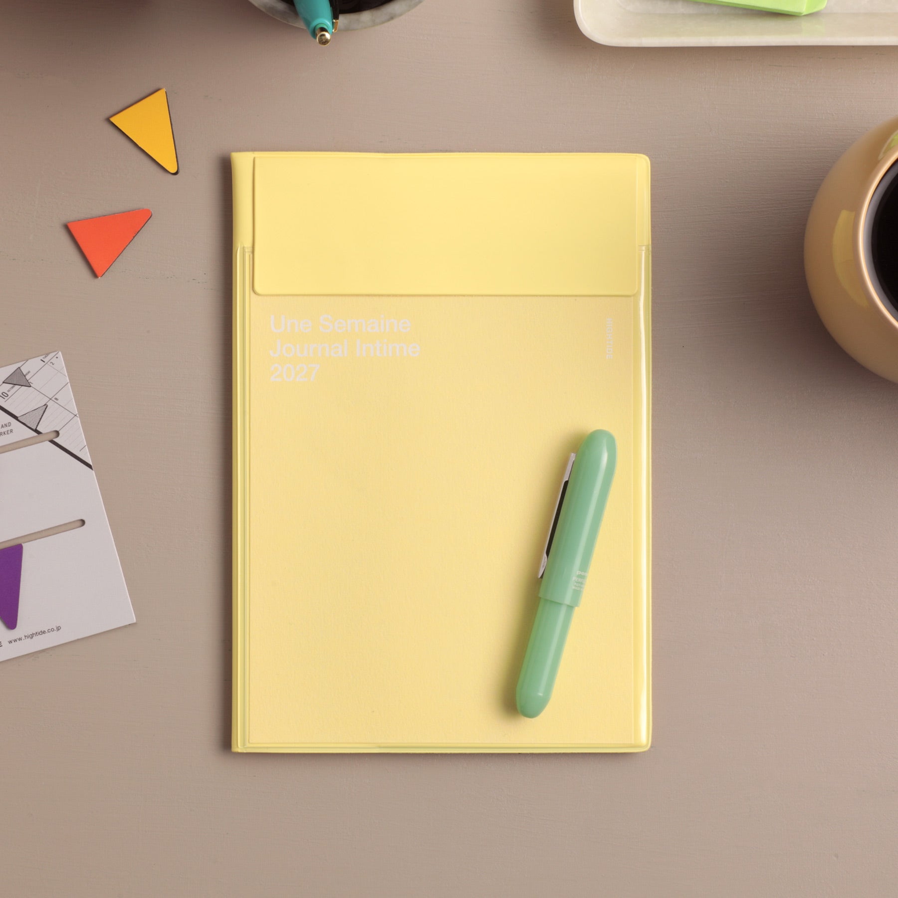 Yellow 2027 monthly planner with envelope-style pocket cover, styled on a desk with a mint pen and triangle clips.