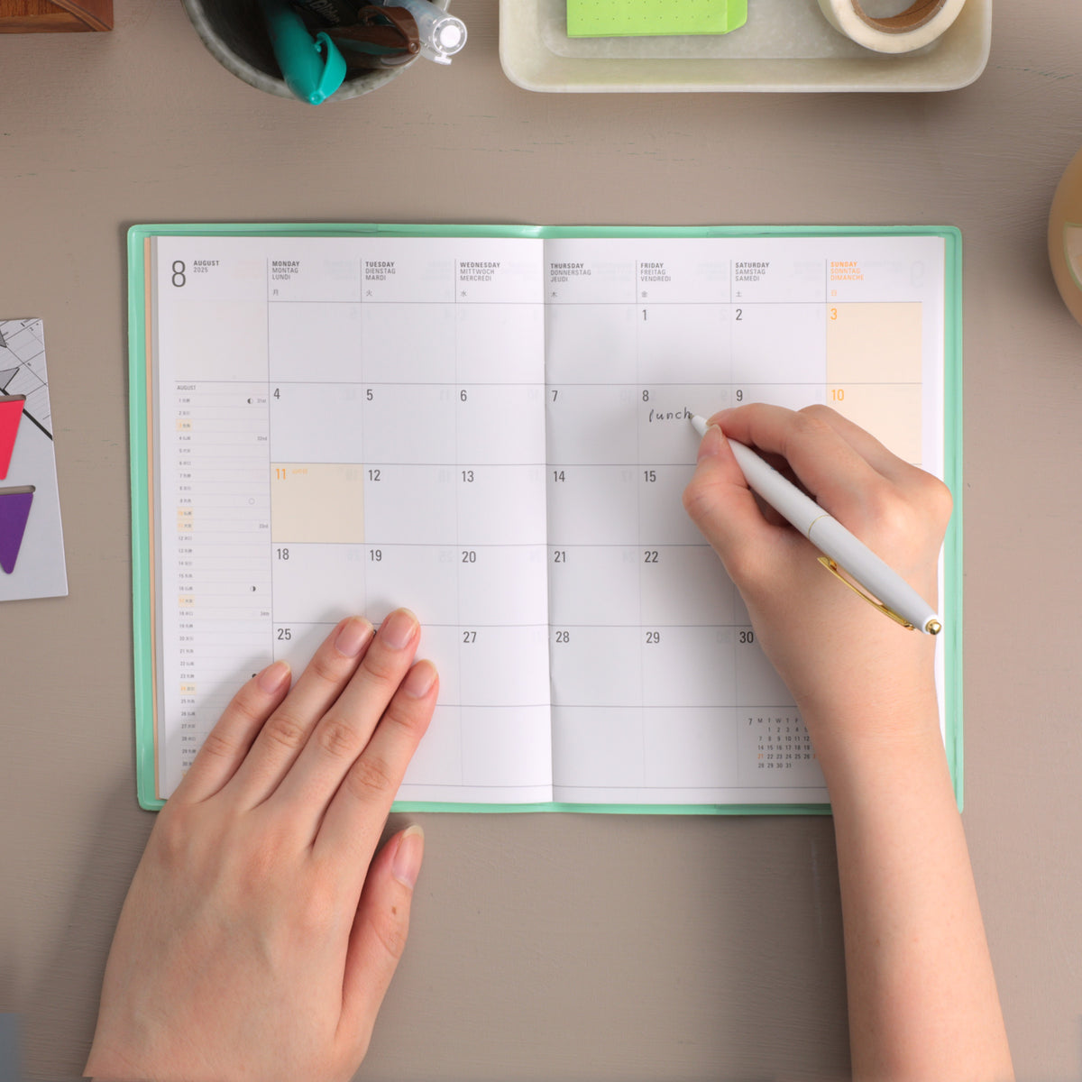 Hands writing on the August monthly grid of an open Mint planner with a pen at a desk.