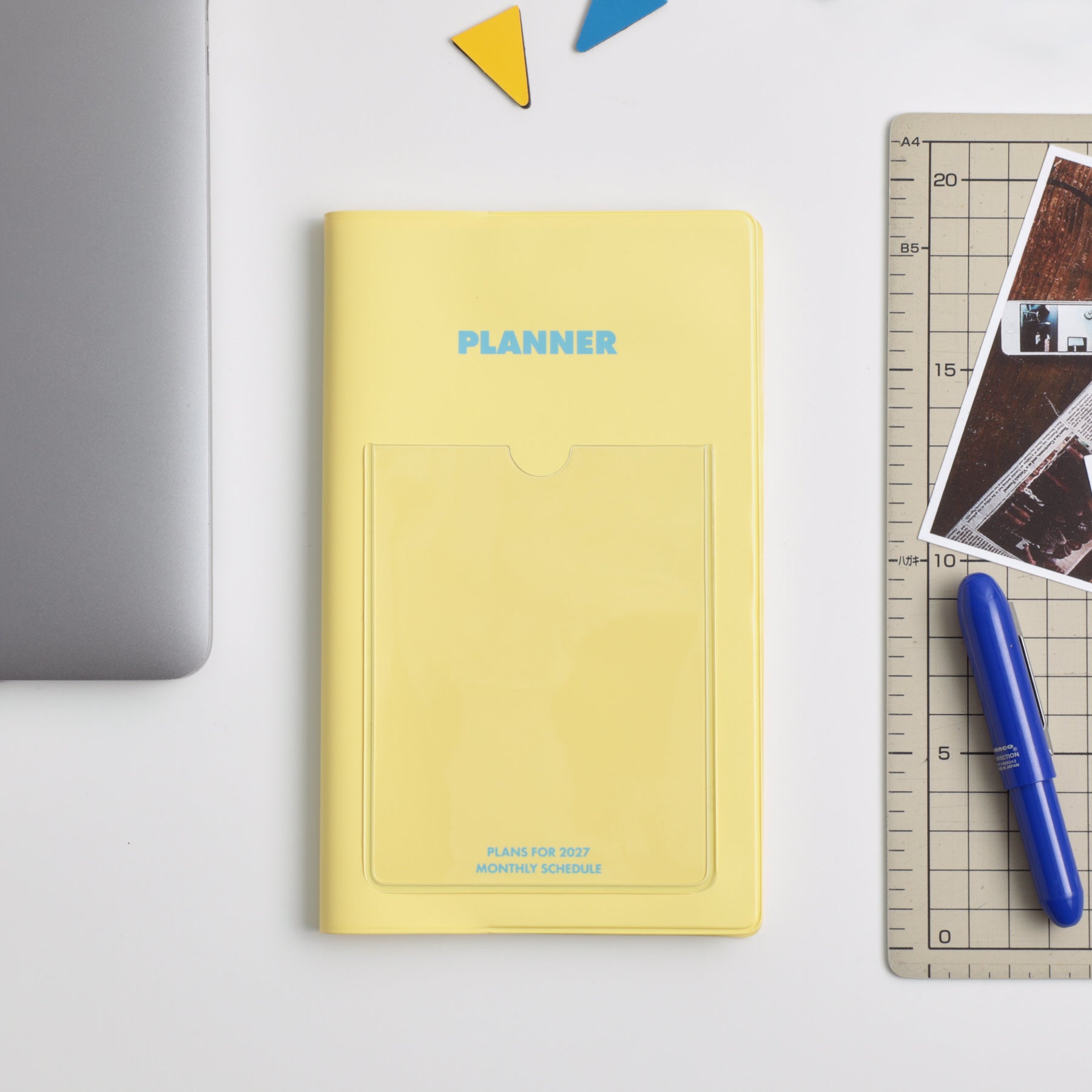 Yellow Hightide Shine planner with front pocket and blue lettering, beside a laptop, ruler, pen and photos.