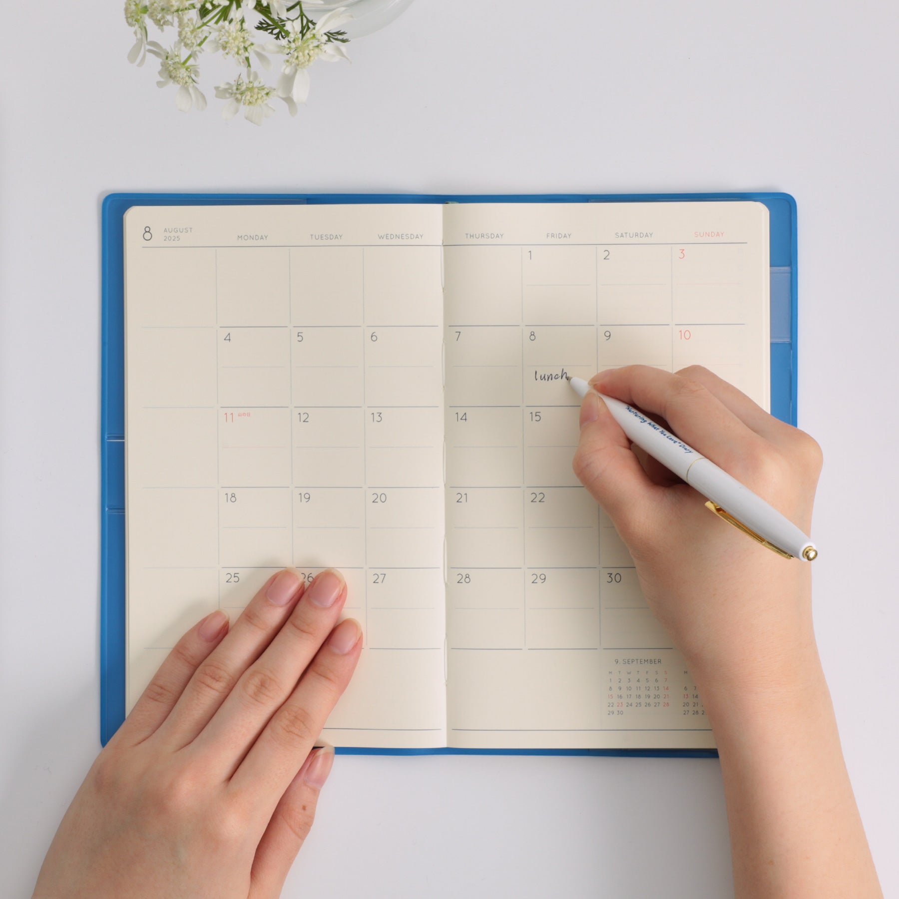 Hands writing in a Blue Hightide planner opened to a monthly calendar spread for August.