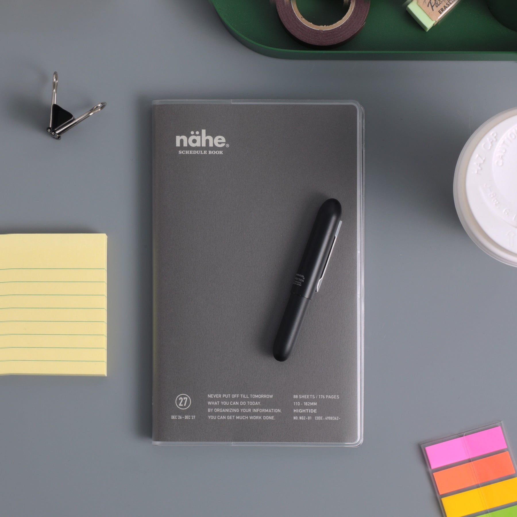 Gray Nähe schedule book in a clear cover with a black pen on top, beside sticky notes and clips.