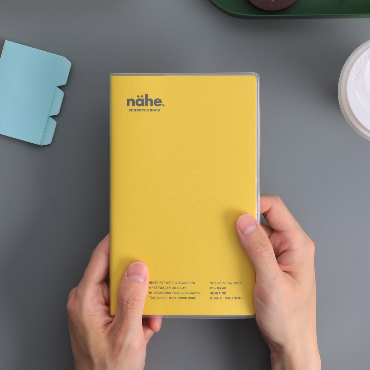 Hands holding a Nähe schedule book in Yellow with a clear cover, over a grey surface.