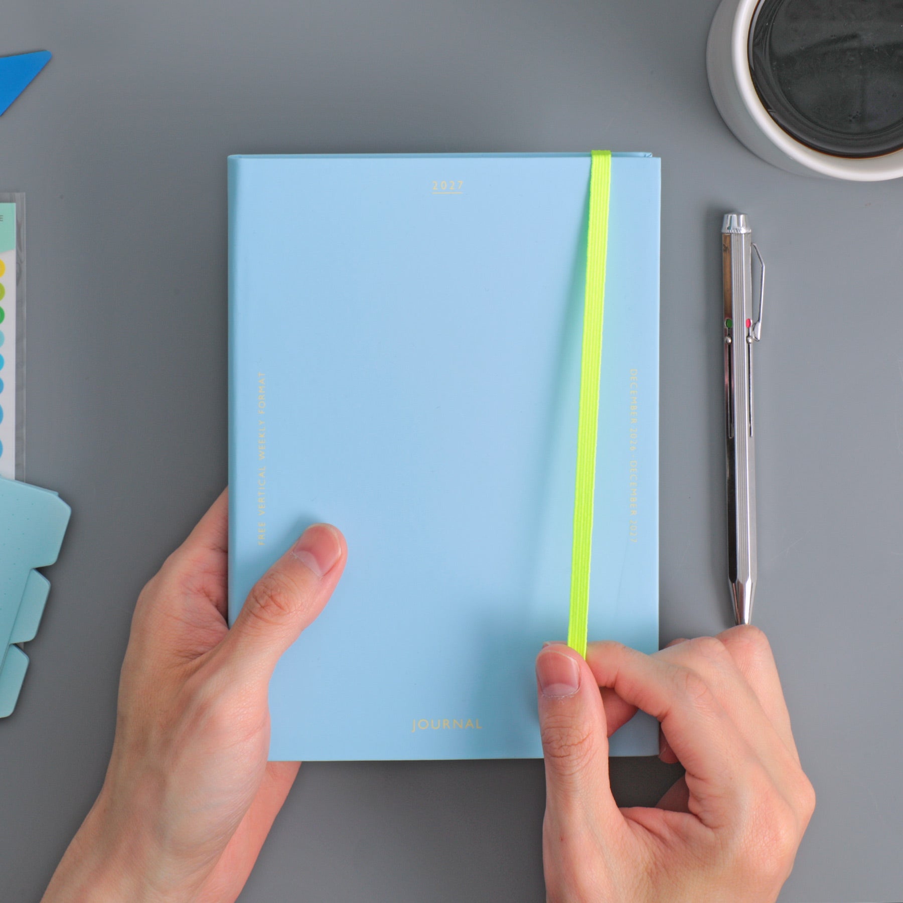 Hands holding a Light Blue Hightide planner with a neon yellow elastic band, beside a pen on a desk.