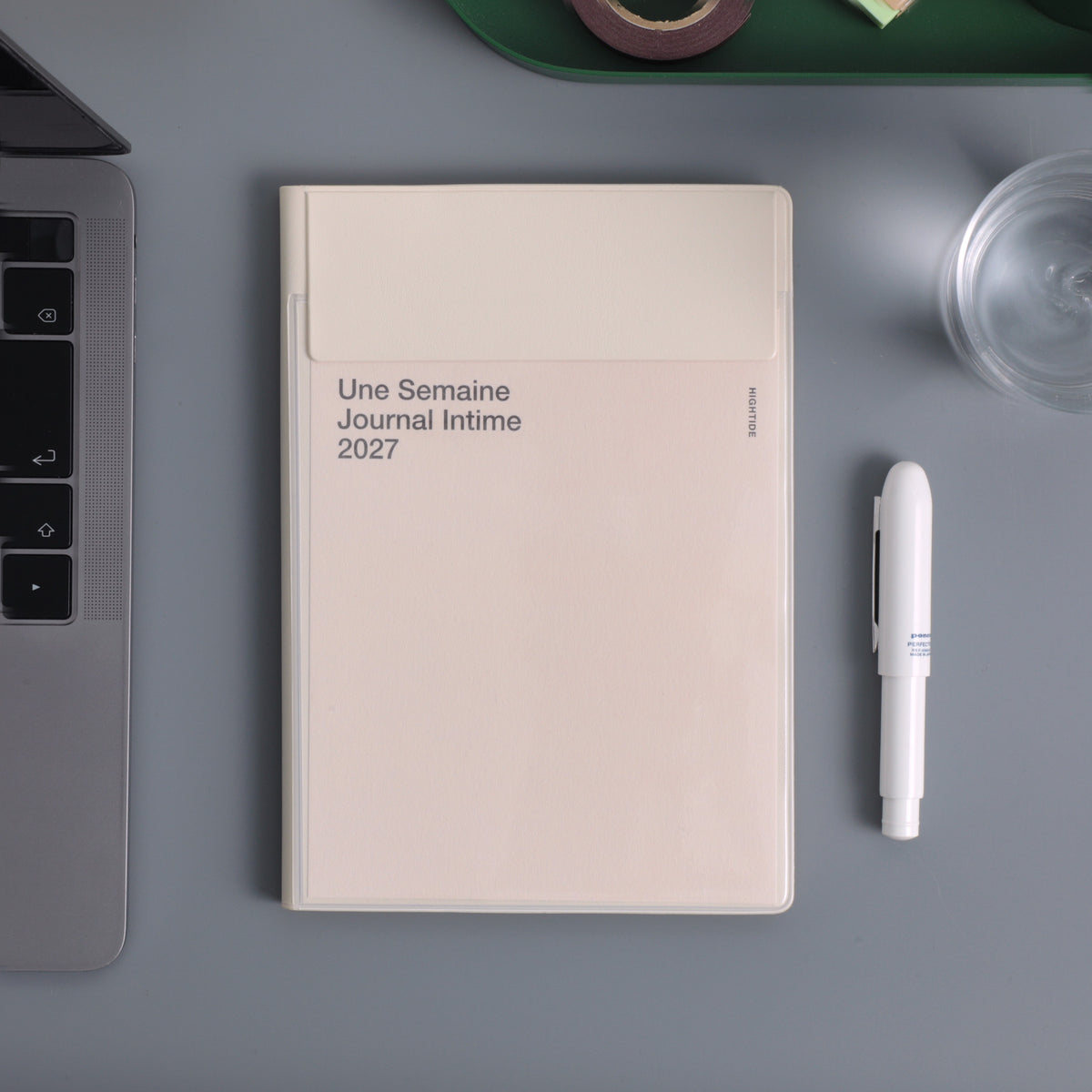 Ivory Hightide 2027 weekly planner with cover pocket, lying on a desk beside a laptop and white pen.