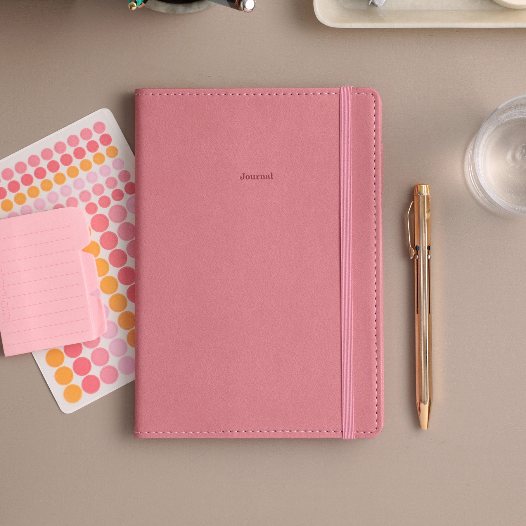 Pink velour weekly planner with matching elastic band and "Journal" text, beside a gold pen and dot stickers.
