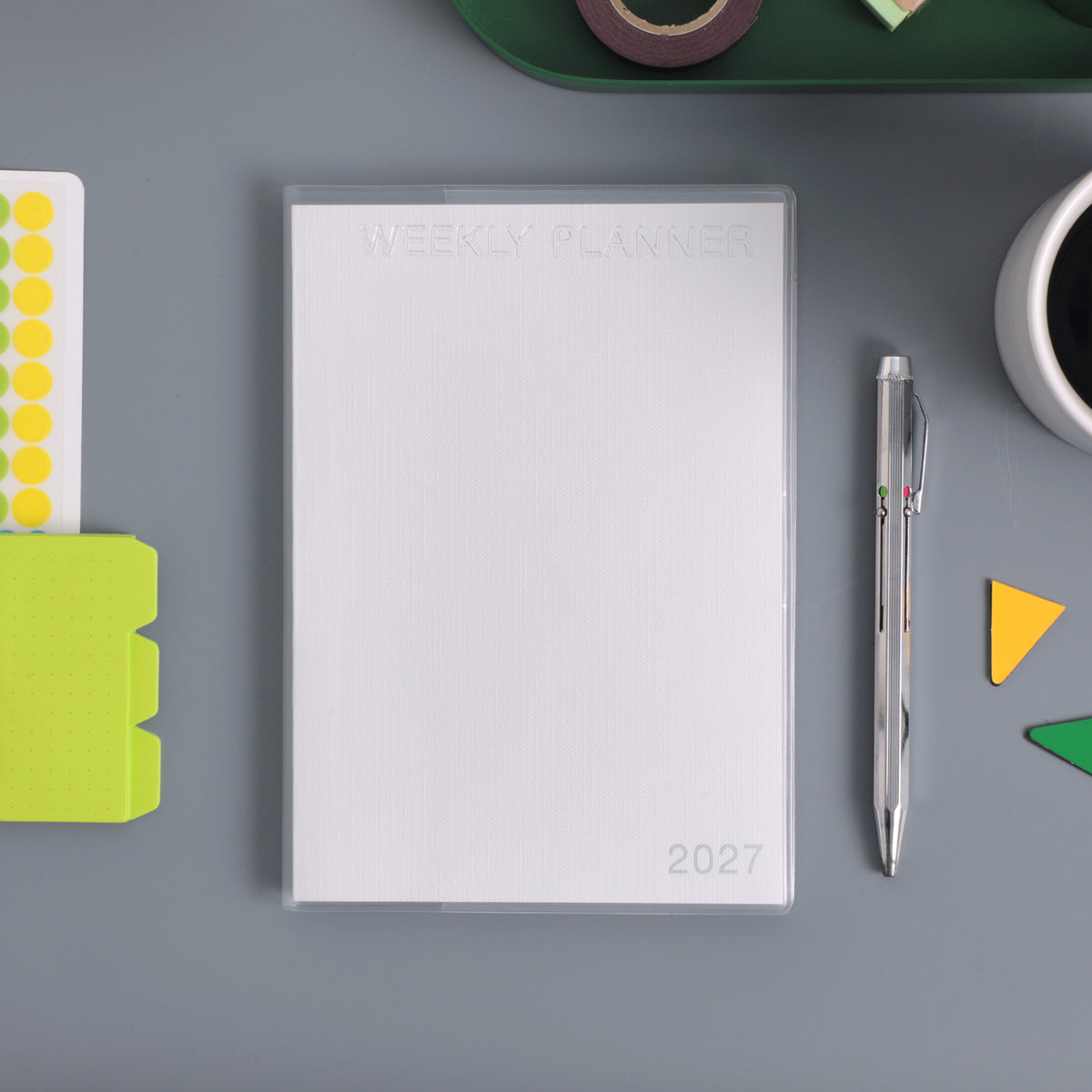 White 2027 Weekly Planner with embossed clear cover, shown flat beside a pen, sticky notes and washi tape.