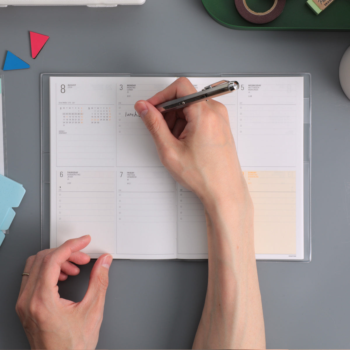 Hands writing with a pen in an open weekly planner with clear cover on a grey desk