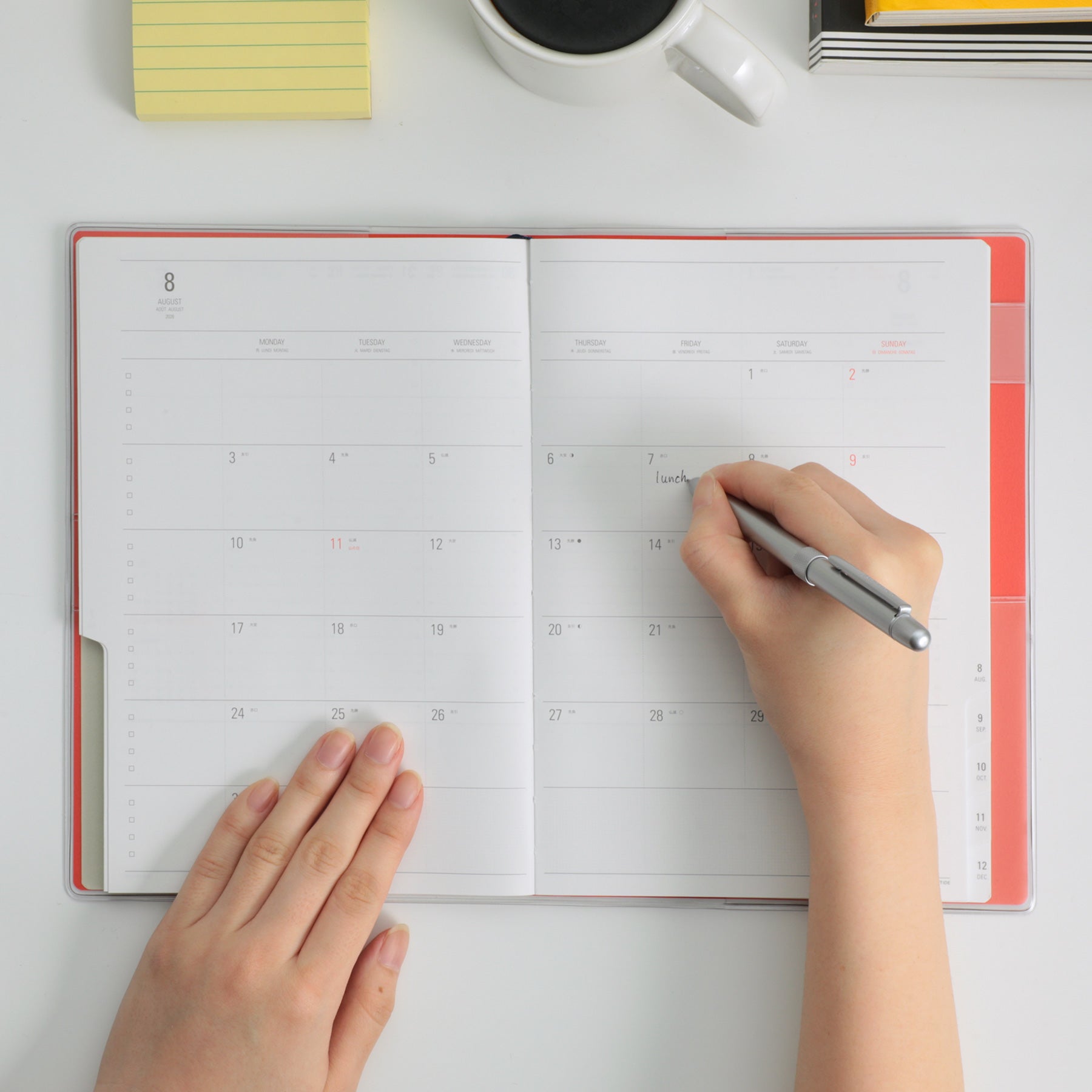 Hands writing with a silver pen in an open Nähe planner showing a monthly calendar spread, Red cover.