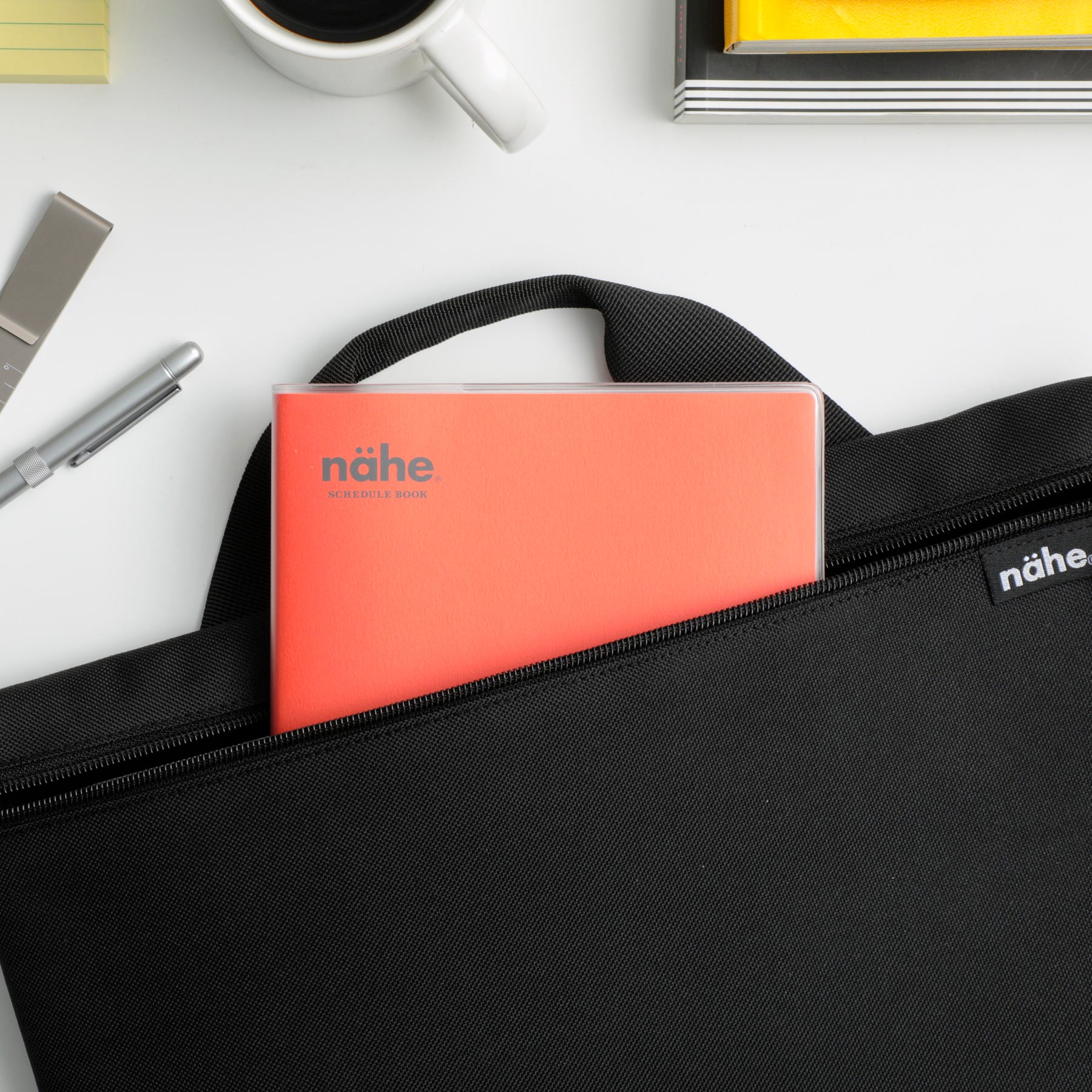 Nähe planner in Red slipped into the front pocket of a black bag, beside a coffee cup and pen.