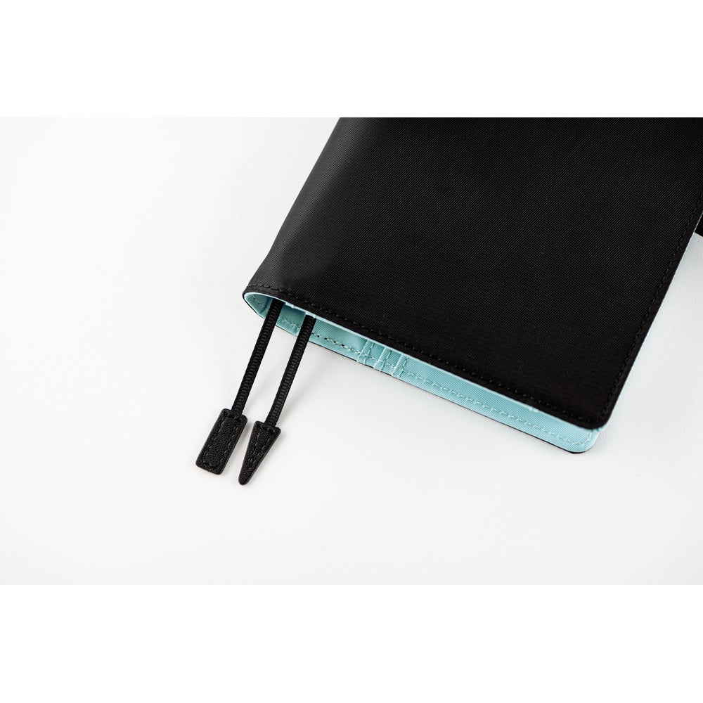 Close-up of a Black A6 book cover corner with two bookmark cords and a pale blue planner inside.