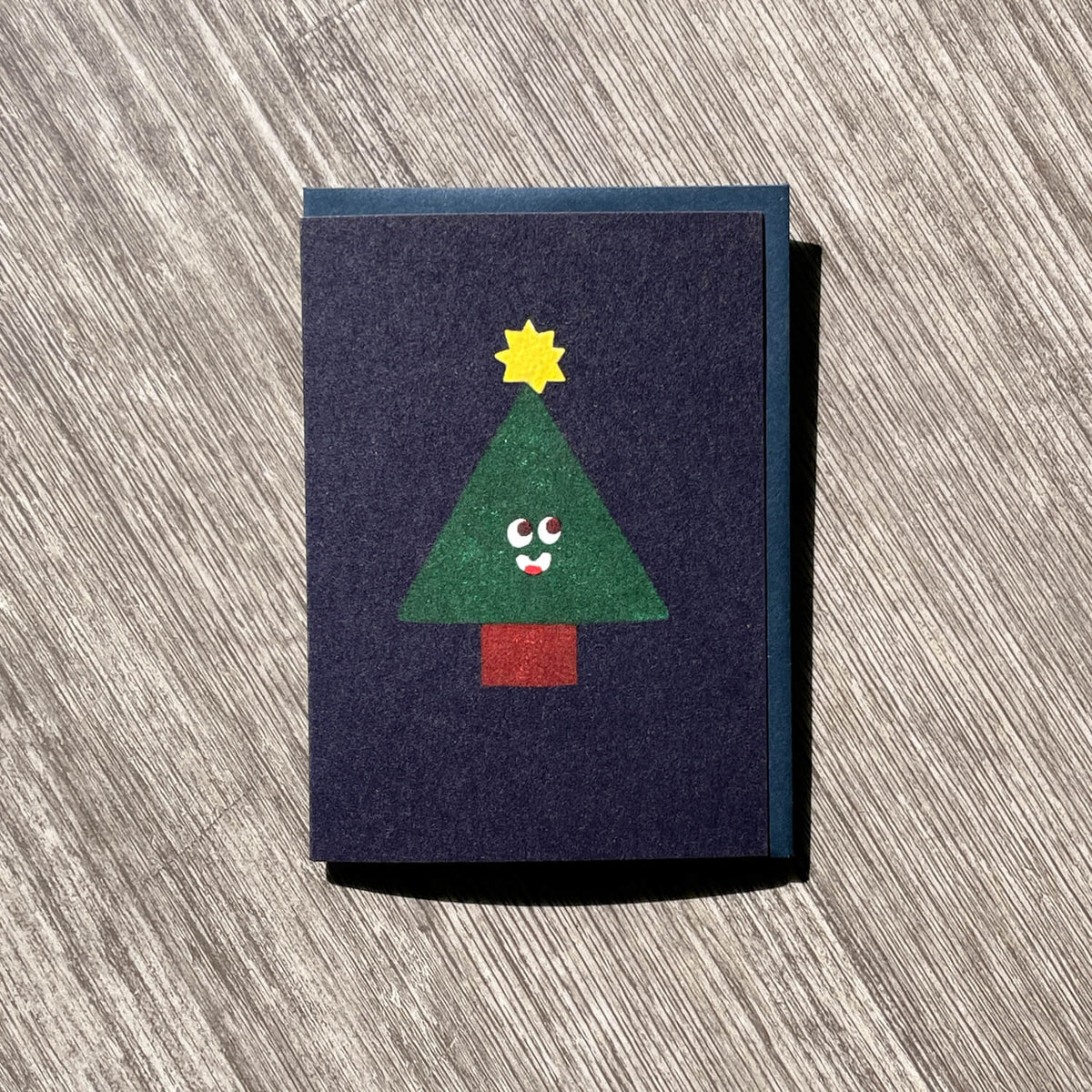 Navy greeting card with a green Christmas tree with a smiling face, yellow star and red base, on wood.