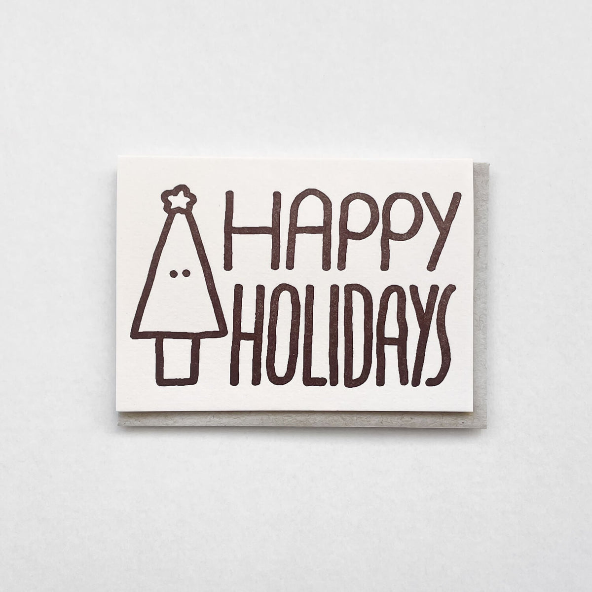Letterpress card with "Happy Holidays" and a smiling fir tree in brown, resting on a grey envelope.