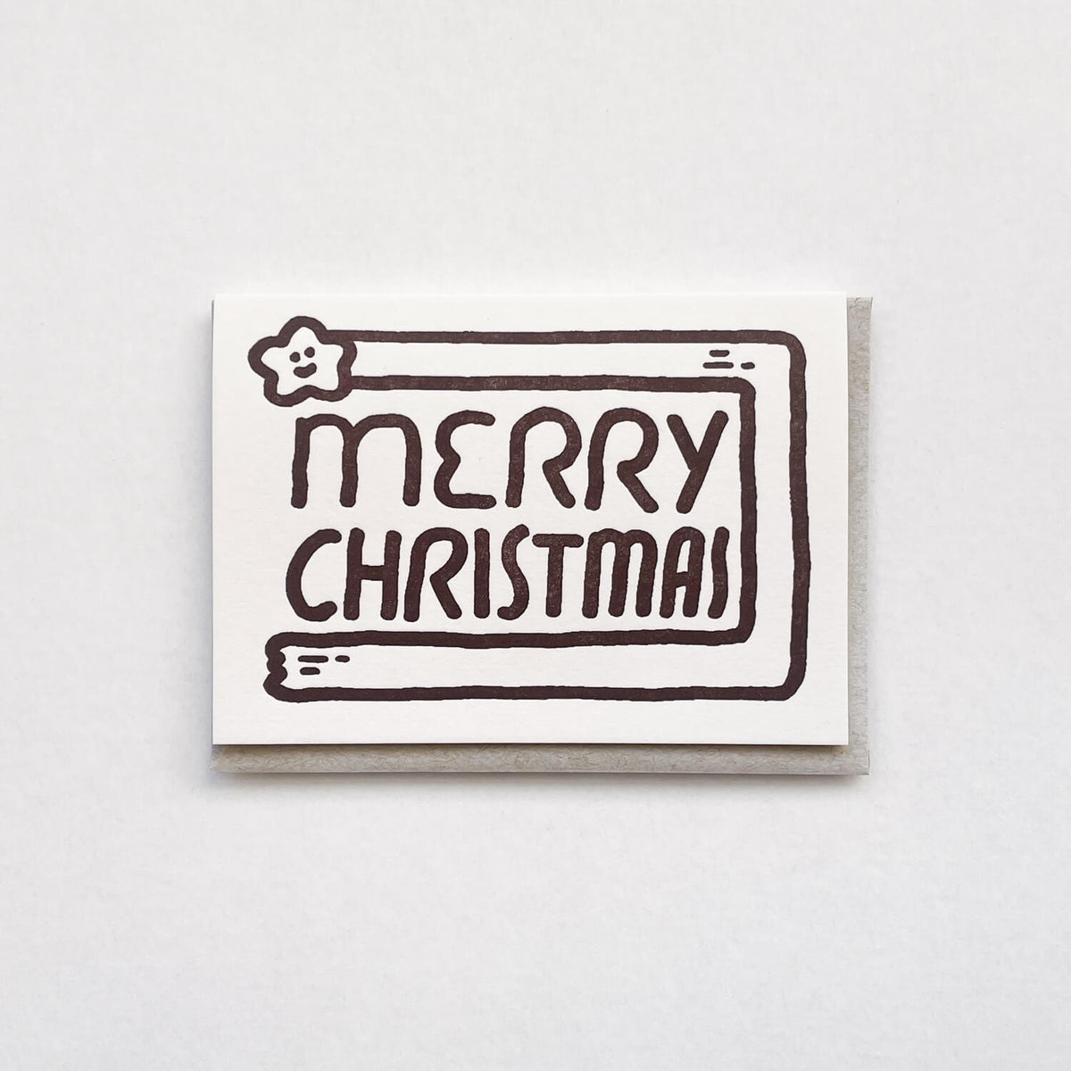 White letterpress card reading "Merry Christmas" in a brown outlined frame with a smiling star, on a grey envelope.
