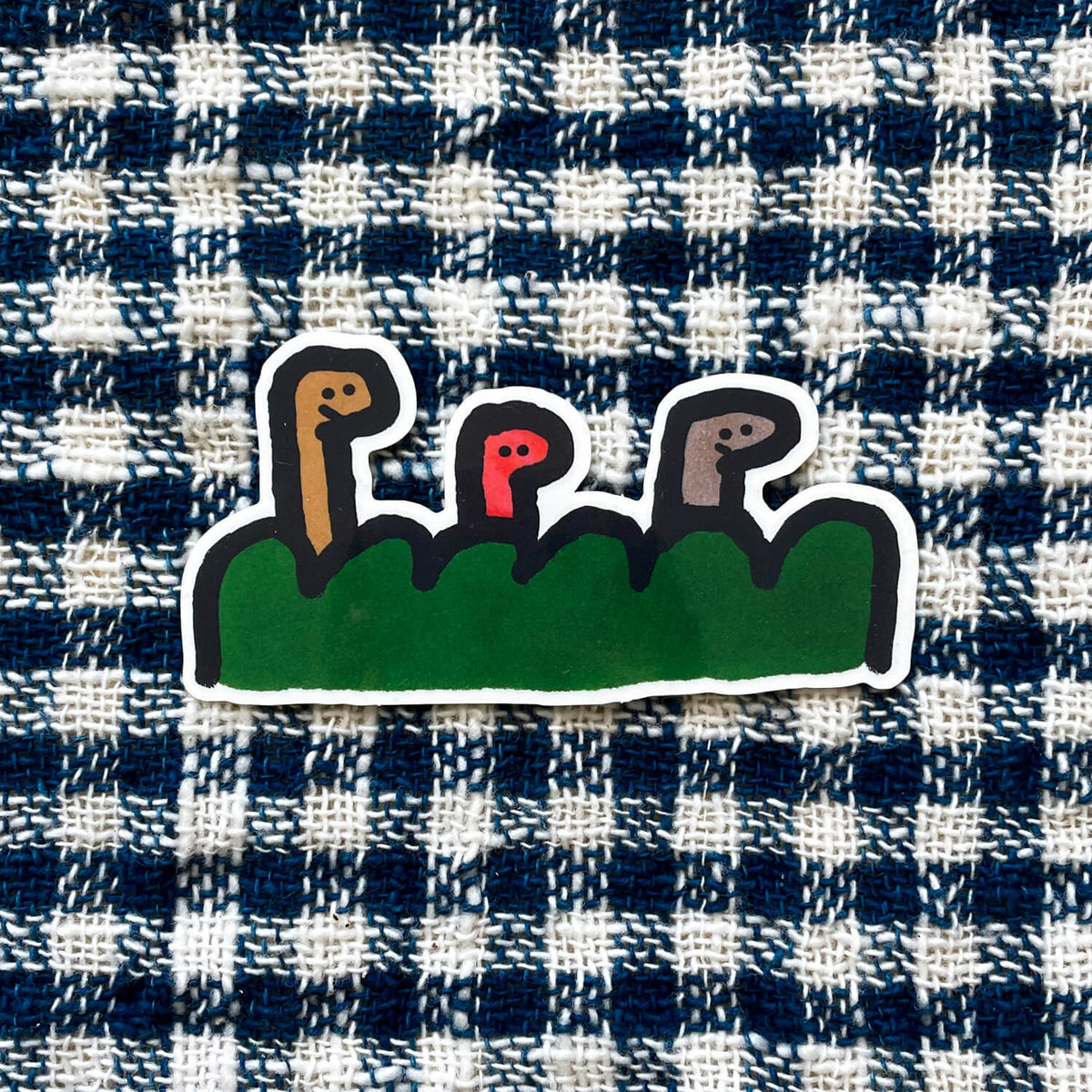 Die-cut sticker of three worm-like figures peeking over green grass, shown on blue checked fabric.