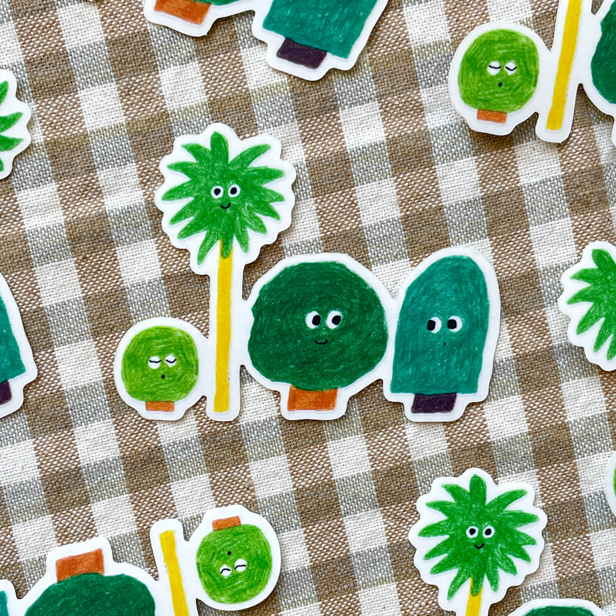 Die-cut stickers of smiling green trees, including a palm and a bush, scattered on beige gingham fabric.