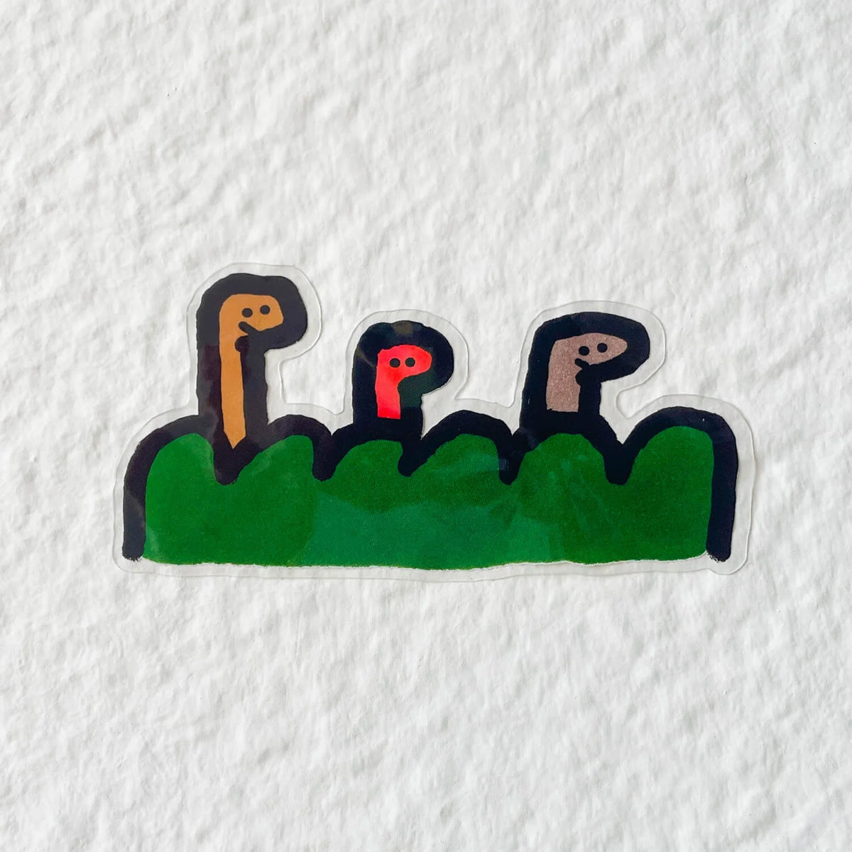 Die-cut clear sticker with three stylized faces peeking over a green bush, on white textured paper.