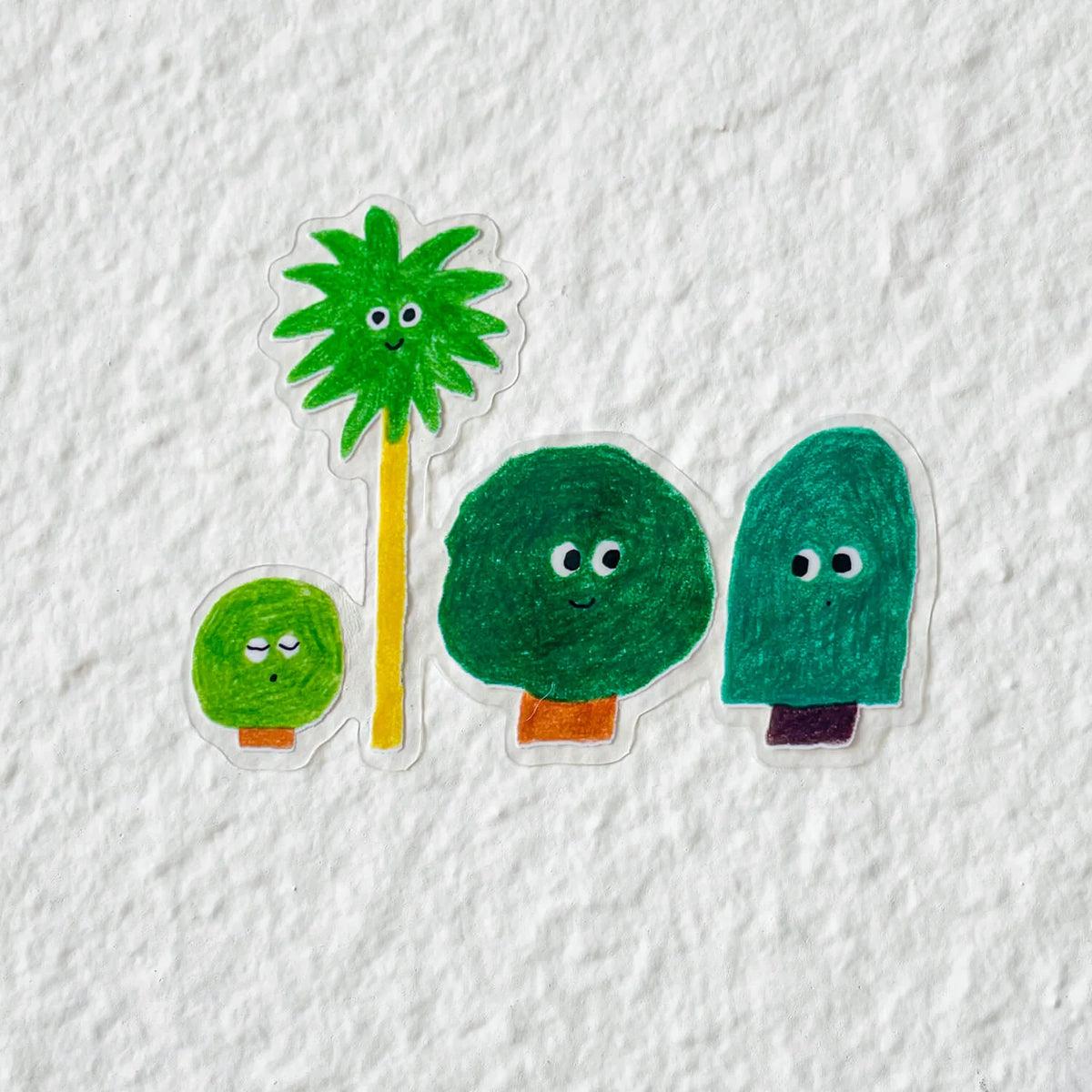 Clear die-cut sticker with four hand-drawn green trees with cartoon faces, on a textured white surface.