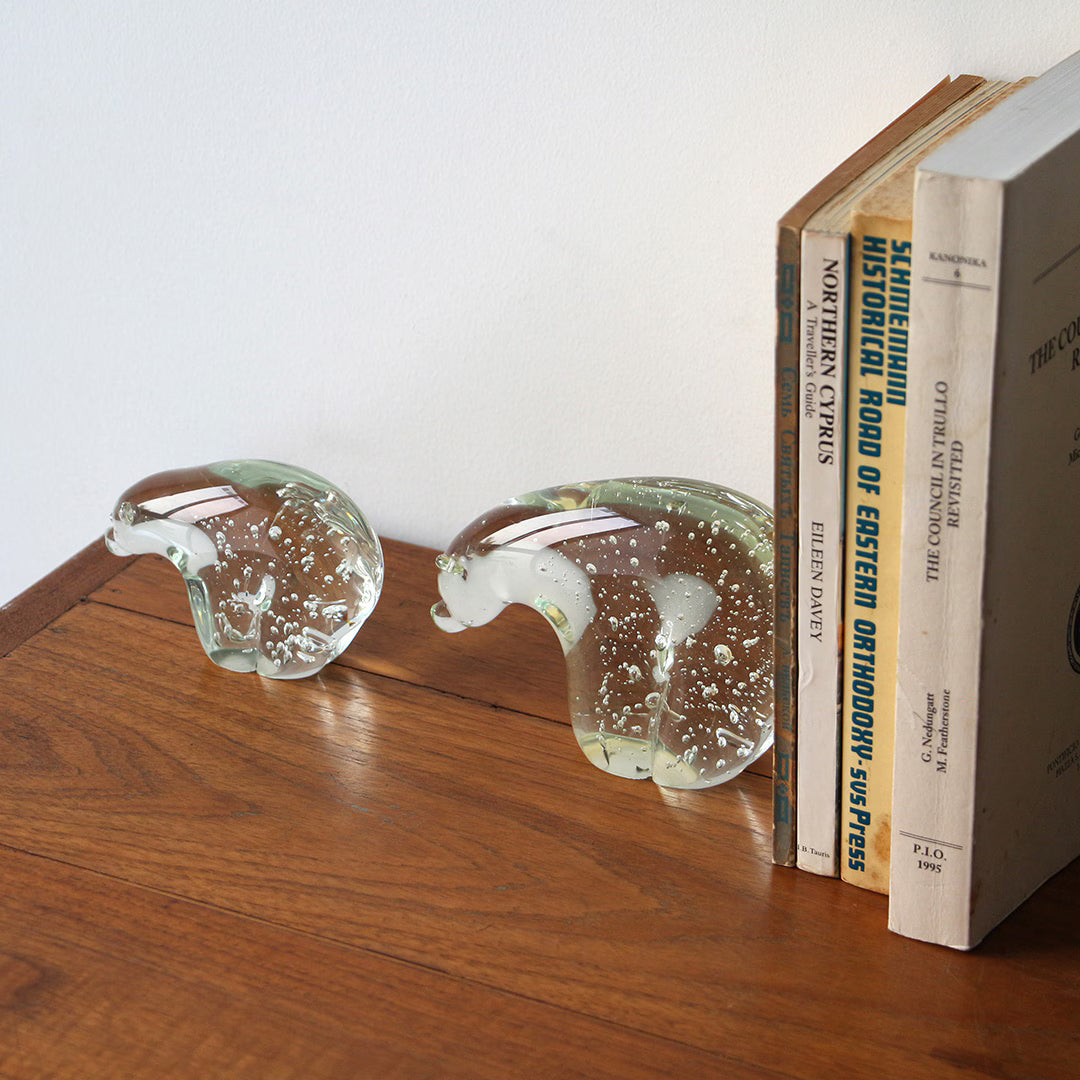 Two clear glass polar bear figures with internal air bubbles on a wooden surface beside leaning books.