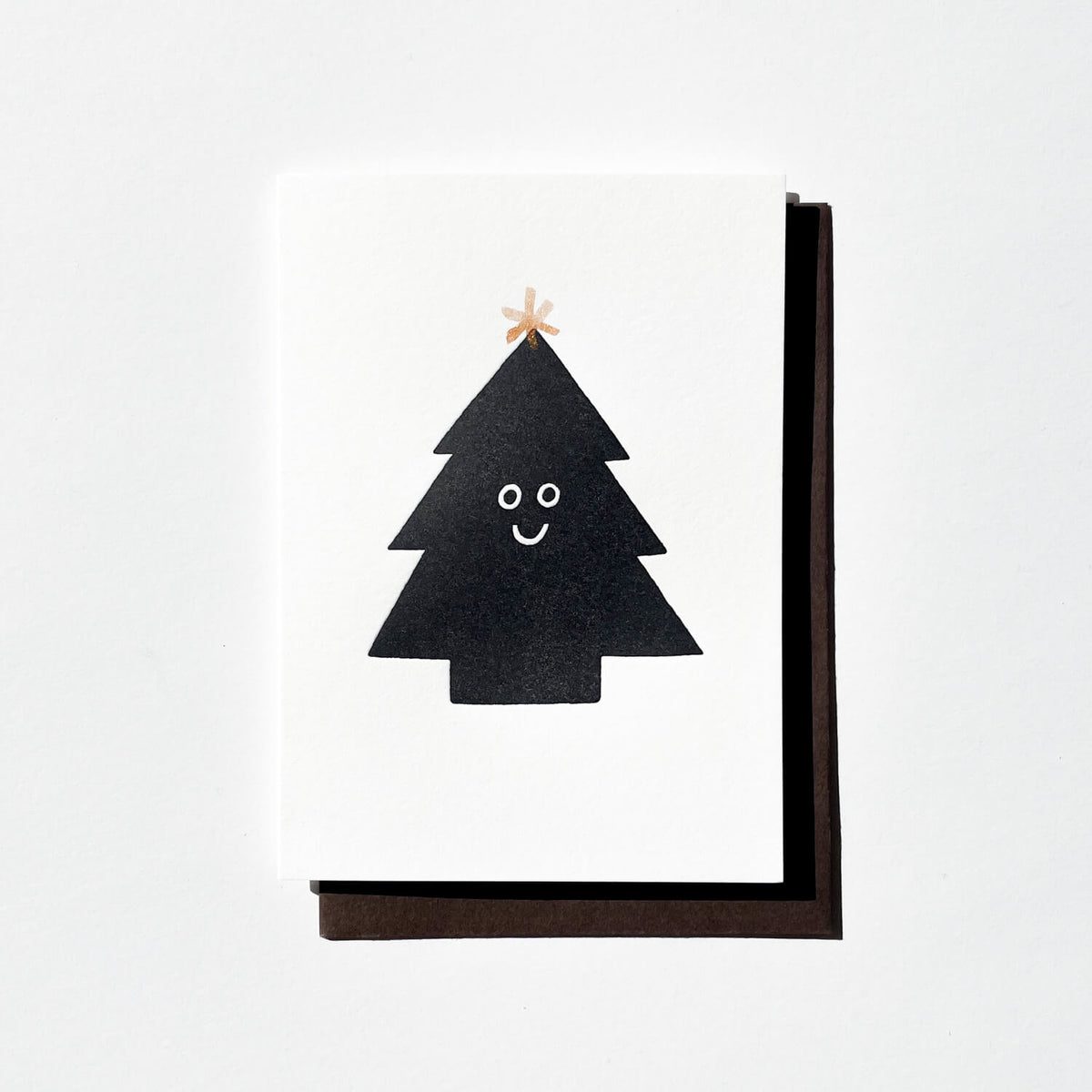 Letterpress card with a smiling black Christmas tree topped by a copper star, with dark envelope behind.
