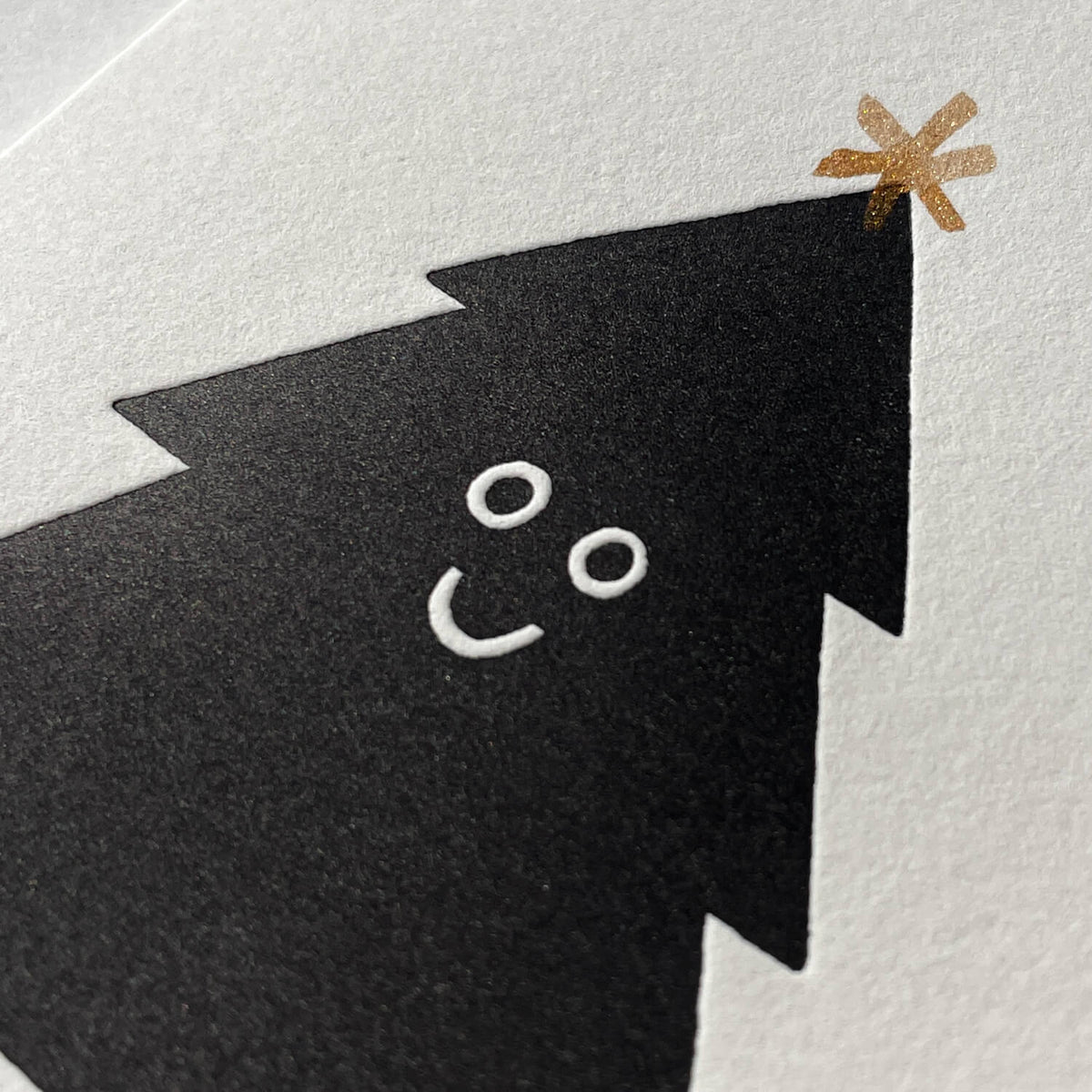 Close-up of letterpress card showing a black tree with a smiling face and gold star on textured paper.
