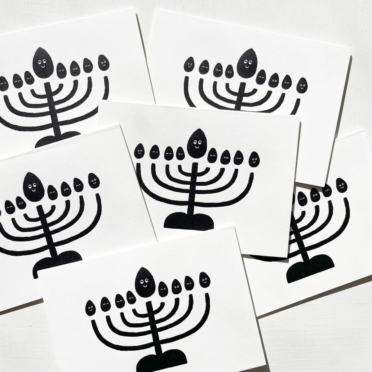 Overlapping white letterpress cards printed with a black menorah whose flames have smiling faces.