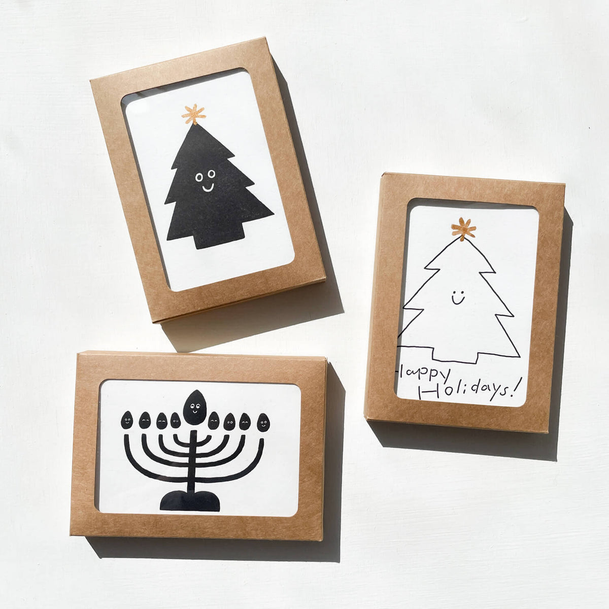 Three kraft boxes with window fronts showing letterpress cards: a black tree, an outlined "Happy Holidays!" tree