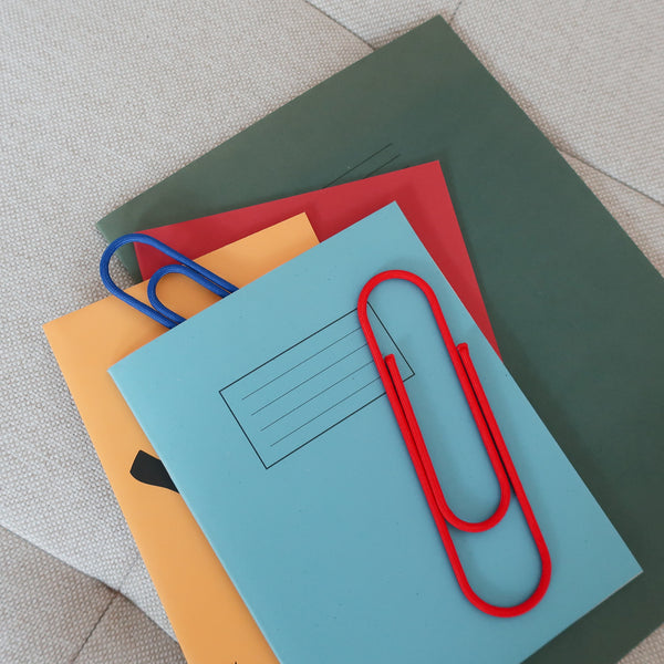 Oversized Red and Blue Onawa clips holding stacked notebooks in blue, orange, red and green.