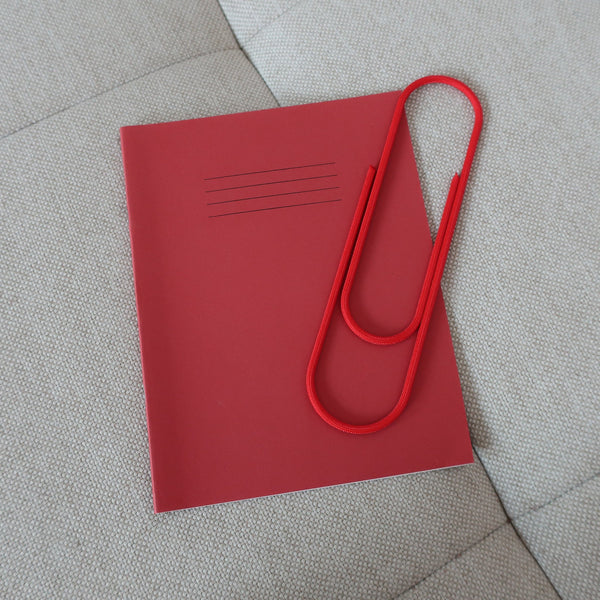Oversized Red cord paper clip resting on a red exercise book, on grey fabric.