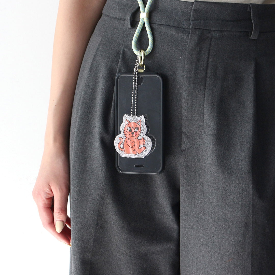 Pink cat reflector keychain hanging from a phone case on a strap, worn against grey trousers.