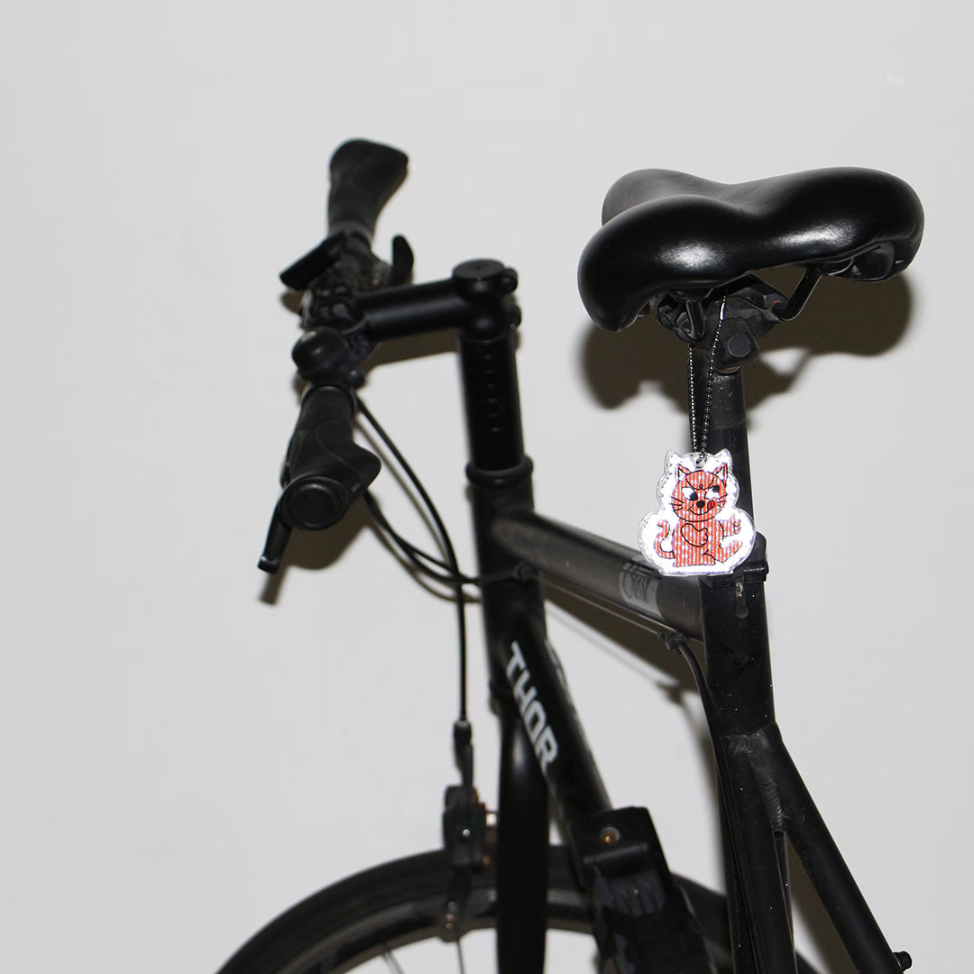 Cheeky cat reflector keychain in Pink hanging by a ball chain from a black bicycle saddle