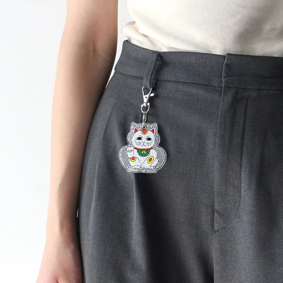 Beckoning cat reflector keychain clipped by its swivel clasp to the belt loop of grey trousers.