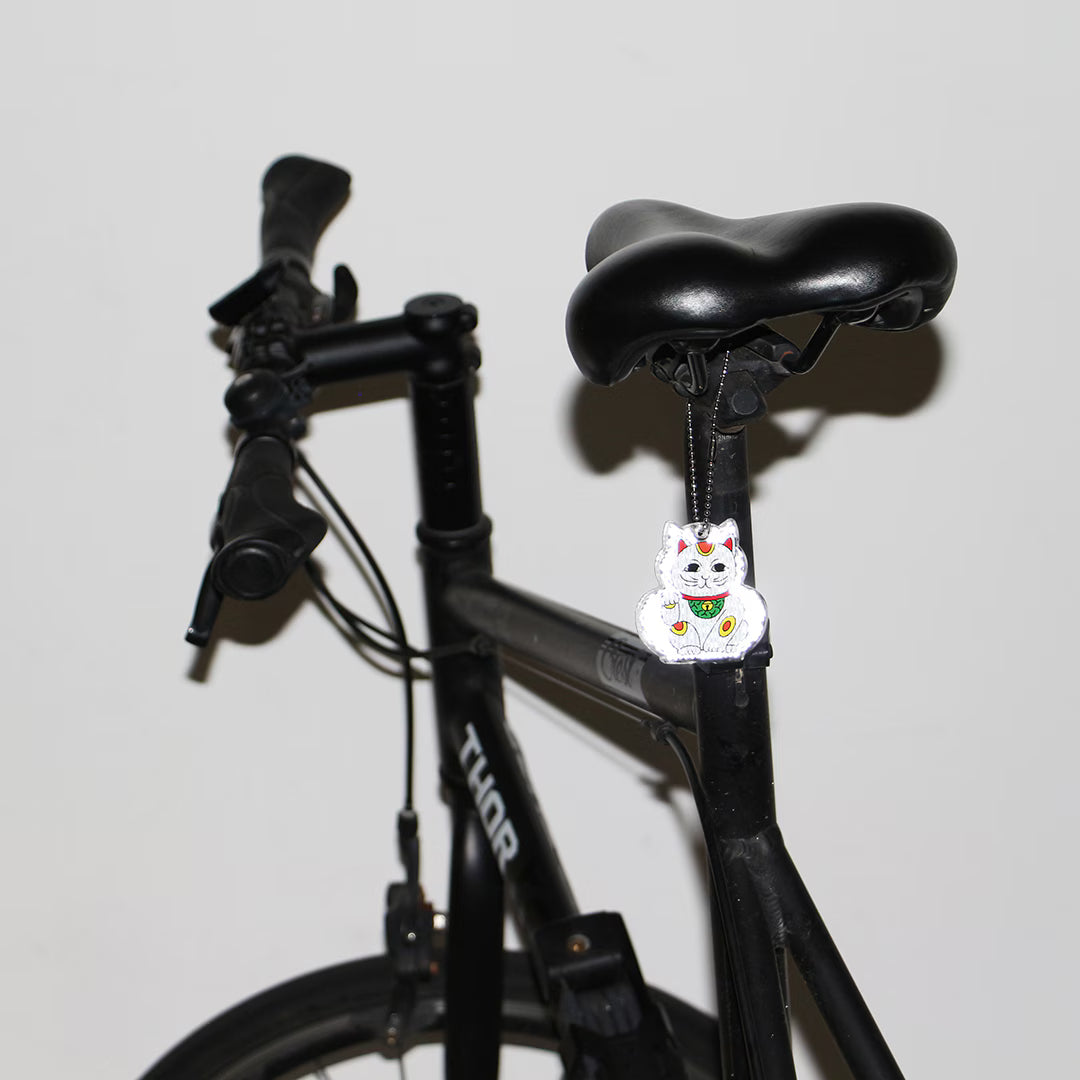 White lucky cat reflector keychain hanging from a black bicycle seat post by a beaded chain.