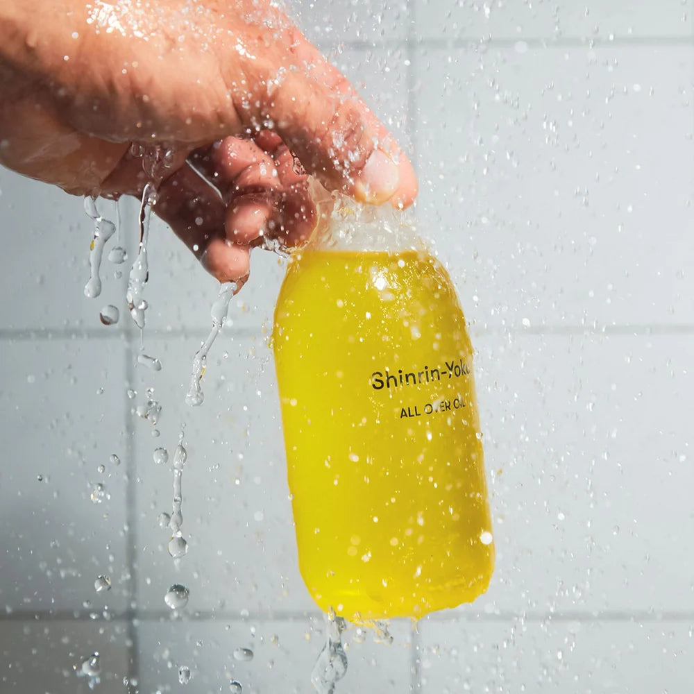 Hand holding a bottle of Earl of East Shinrin-Yoku All Over Oil under running water in a tiled shower.