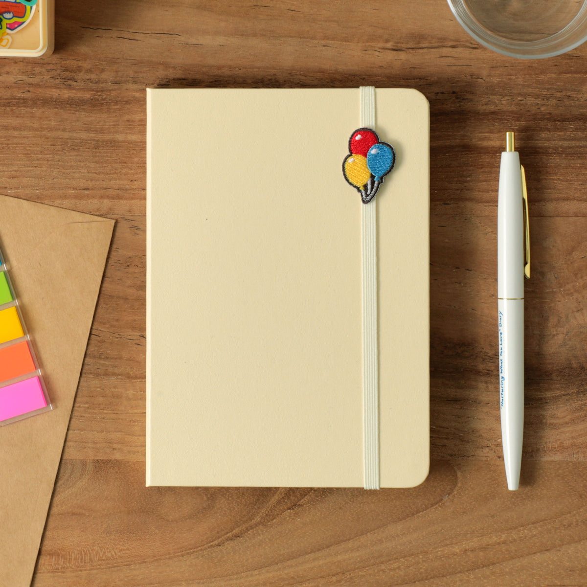 Ivory hardcover A6 planner with elastic band holding an embroidered balloon charm, on a wooden desk with pen.