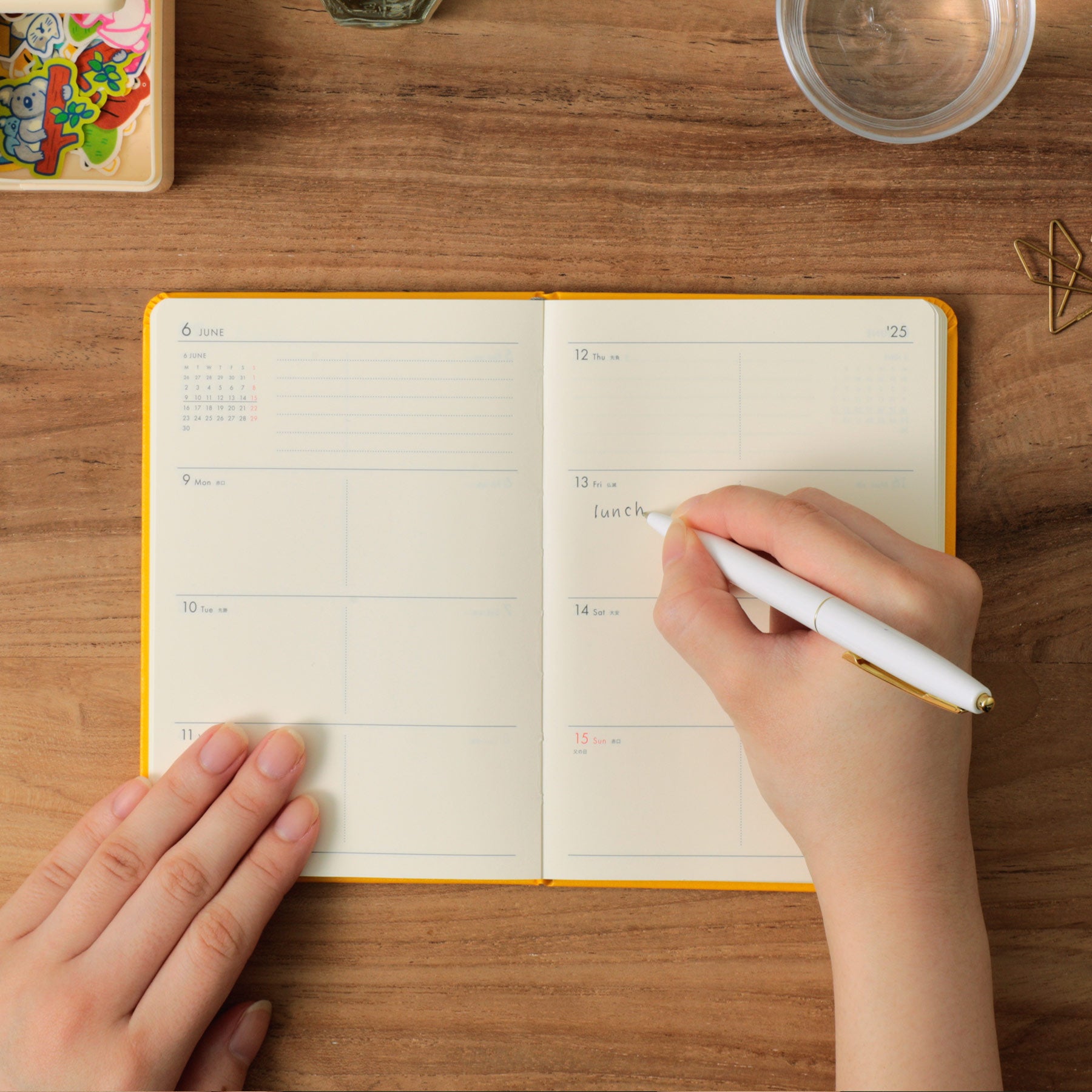 Hands writing "lunch" with a white pen in an open Yellow weekly planner on a wooden desk.