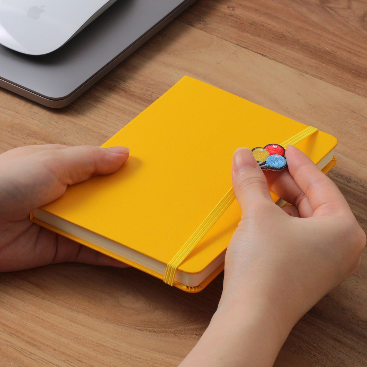 Hands holding a Yellow A6 planner on a wooden desk, sliding the embroidered balloon charm along its elastic band.