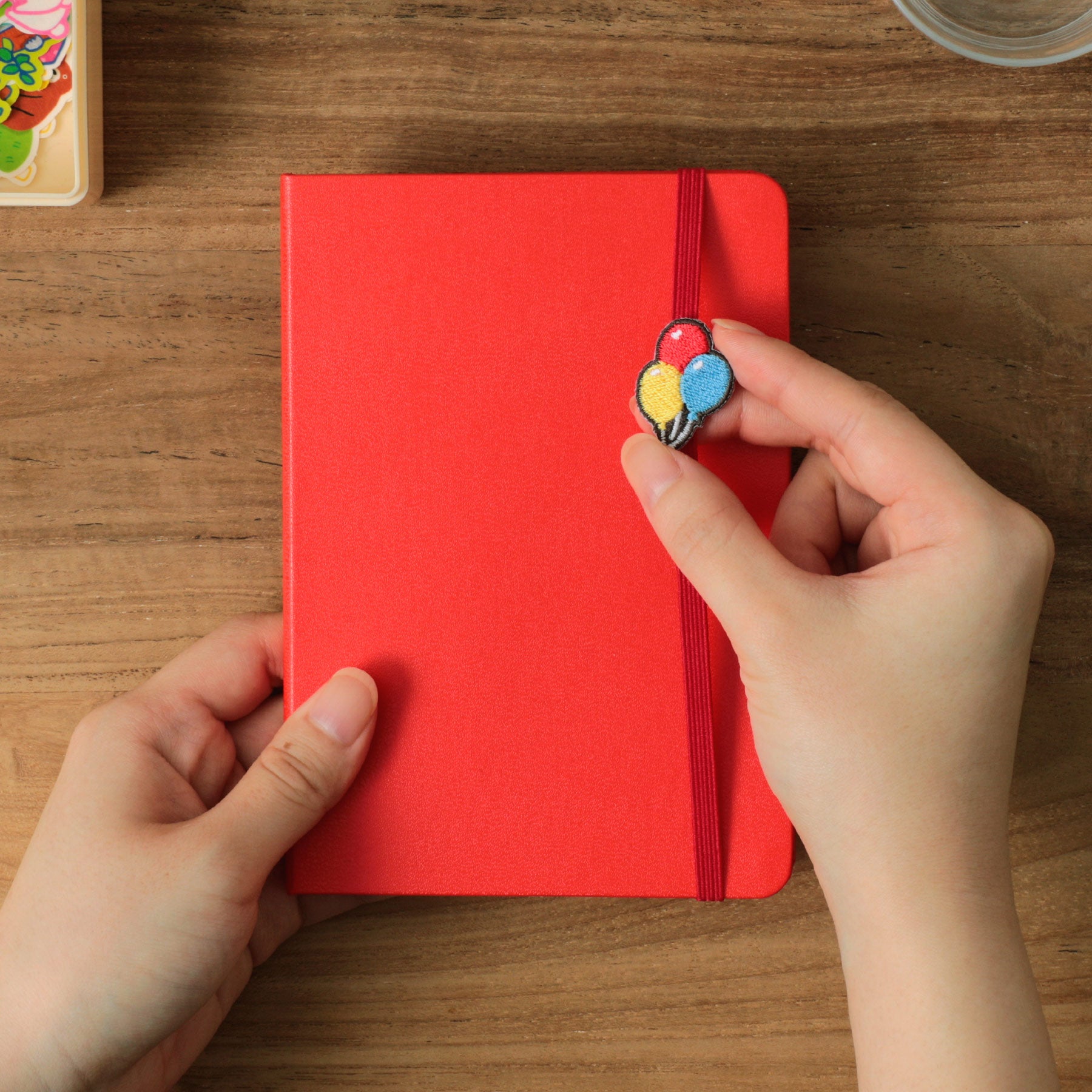 Hands holding a Red hardcover planner on wood, sliding an embroidered balloon charm along the elastic band.