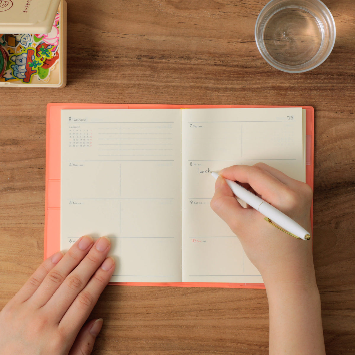 Hands writing "lunch" in an open Pink weekly planner on a wooden table, with stickers and a glass nearby.