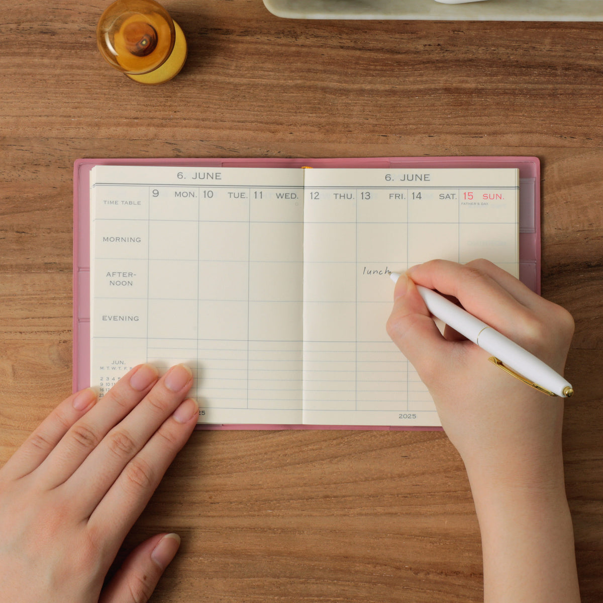Hands writing "lunch" with a white pen in a Pink Hightide planner open to a weekly June spread on wood.
